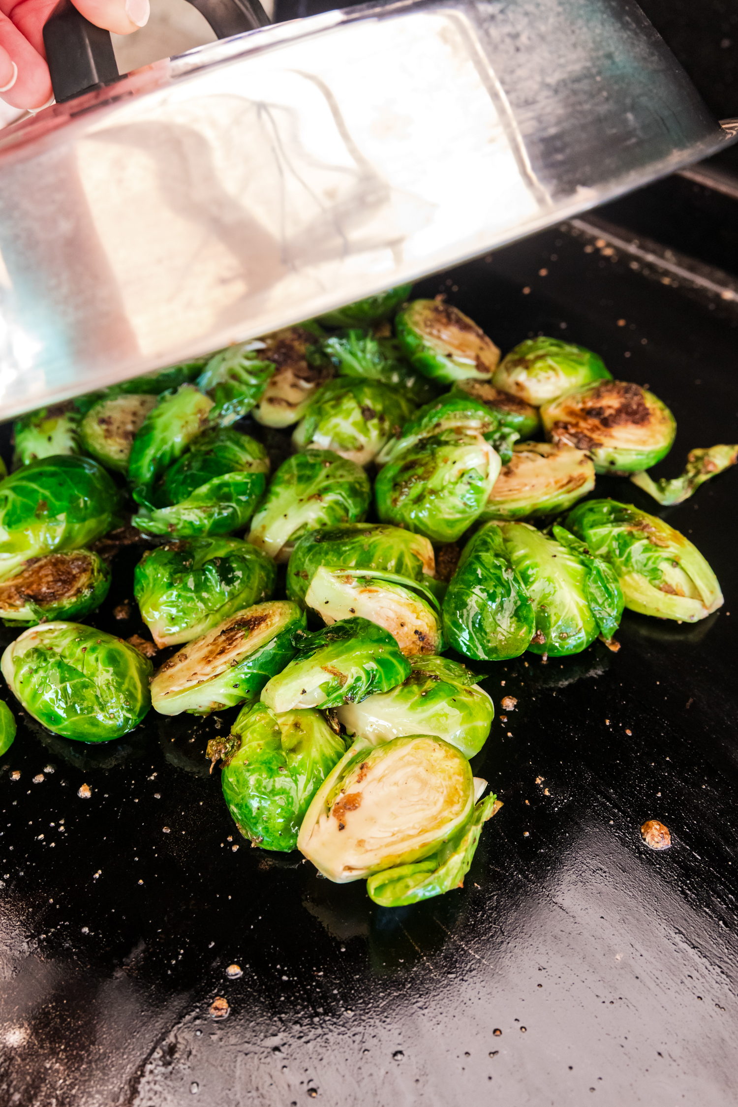 A hand holding a lid over Brussels sprouts on a griddle, allowing them to steam while cooking.