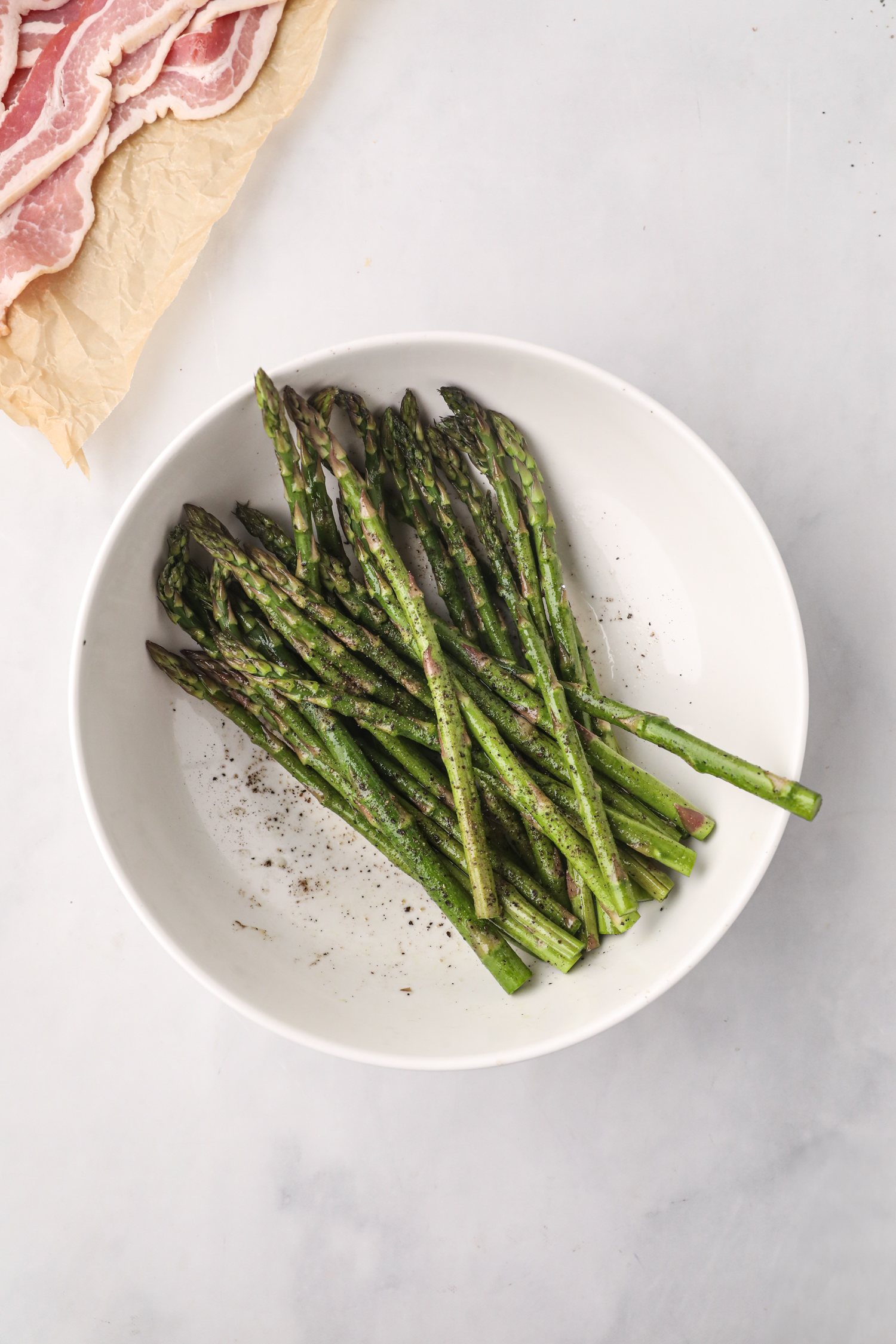 Fresh asparagus spears seasoned with salt and pepper in a white bowl, with slices of raw bacon visible on parchment paper beside it.