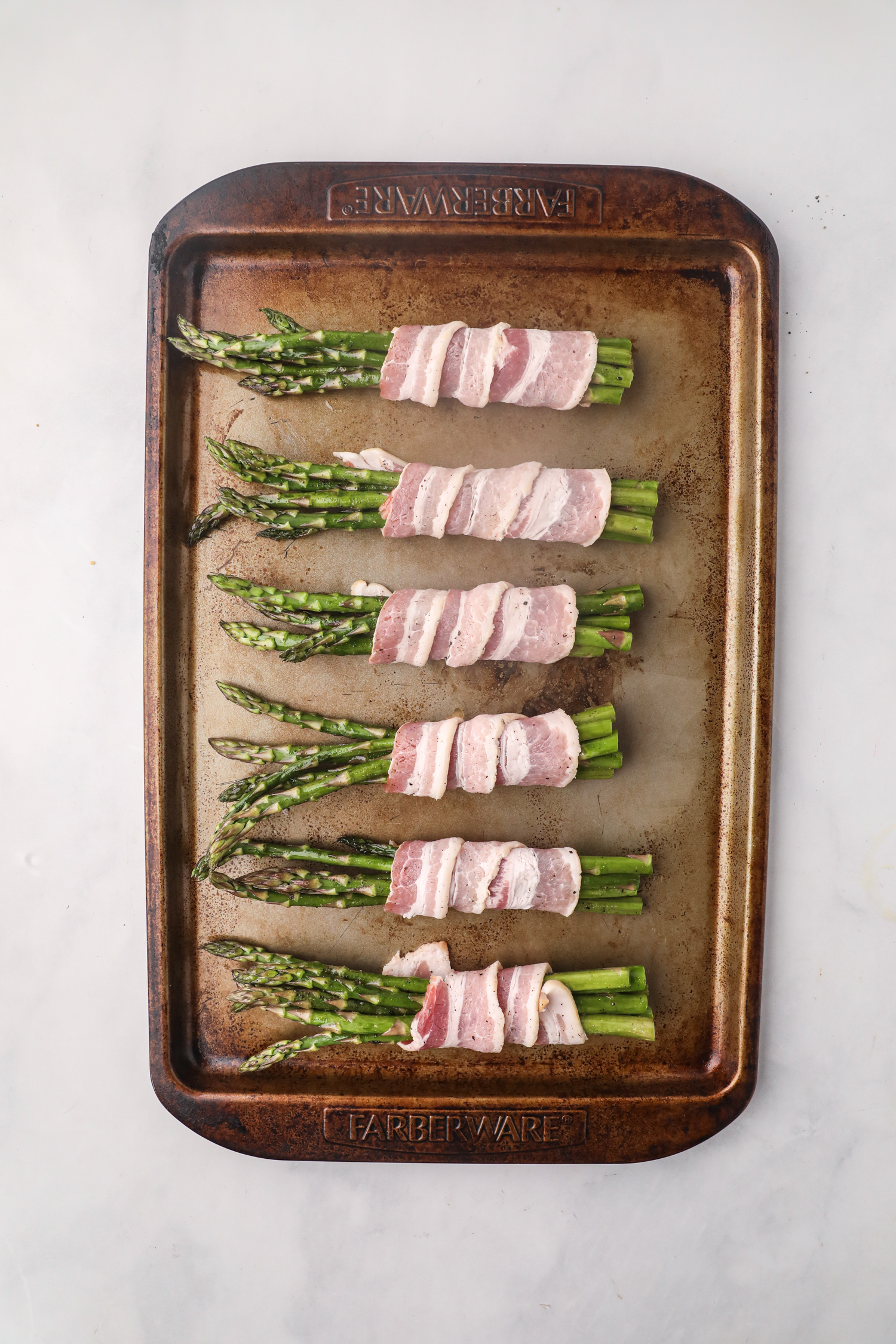 Bundles of asparagus spears wrapped in raw bacon arranged evenly on a baking sheet, ready for grilling.