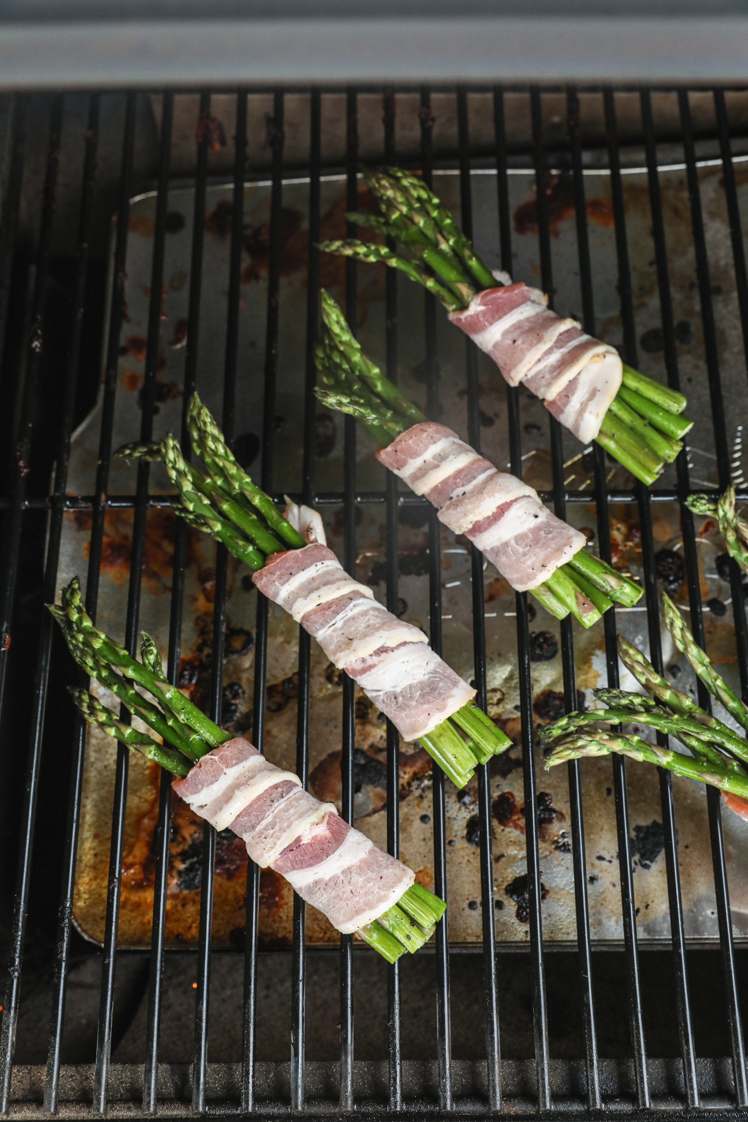 Asparagus bundles wrapped in raw bacon placed on a grill grate above a drip tray, beginning the cooking process.