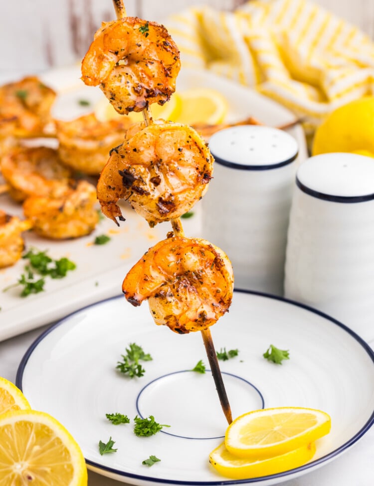 A close-up of a grilled shrimp skewer held upright over a white plate with blue trim, garnished with parsley and lemon slices; the background shows more shrimp and salt and pepper shakers.