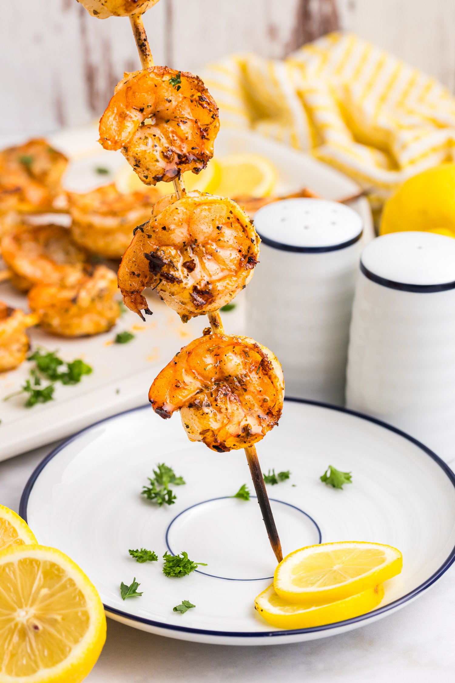 A close-up of a grilled shrimp skewer held upright over a white plate with blue trim, garnished with parsley and lemon slices; the background shows more shrimp and salt and pepper shakers.

