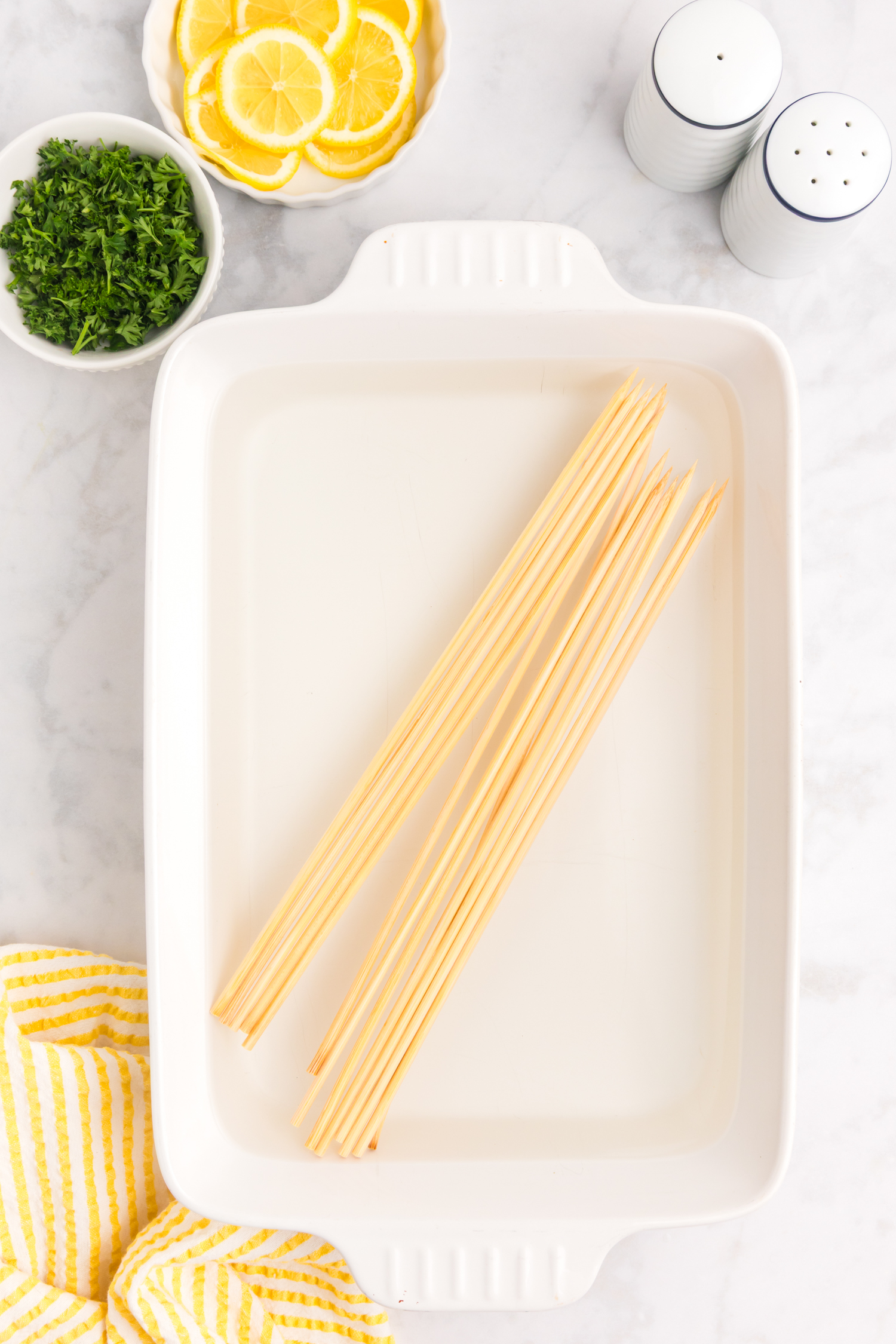 A white rectangular baking dish with several wooden skewers laid inside it, surrounded by small bowls of lemon slices and chopped parsley, with salt and pepper shakers nearby.