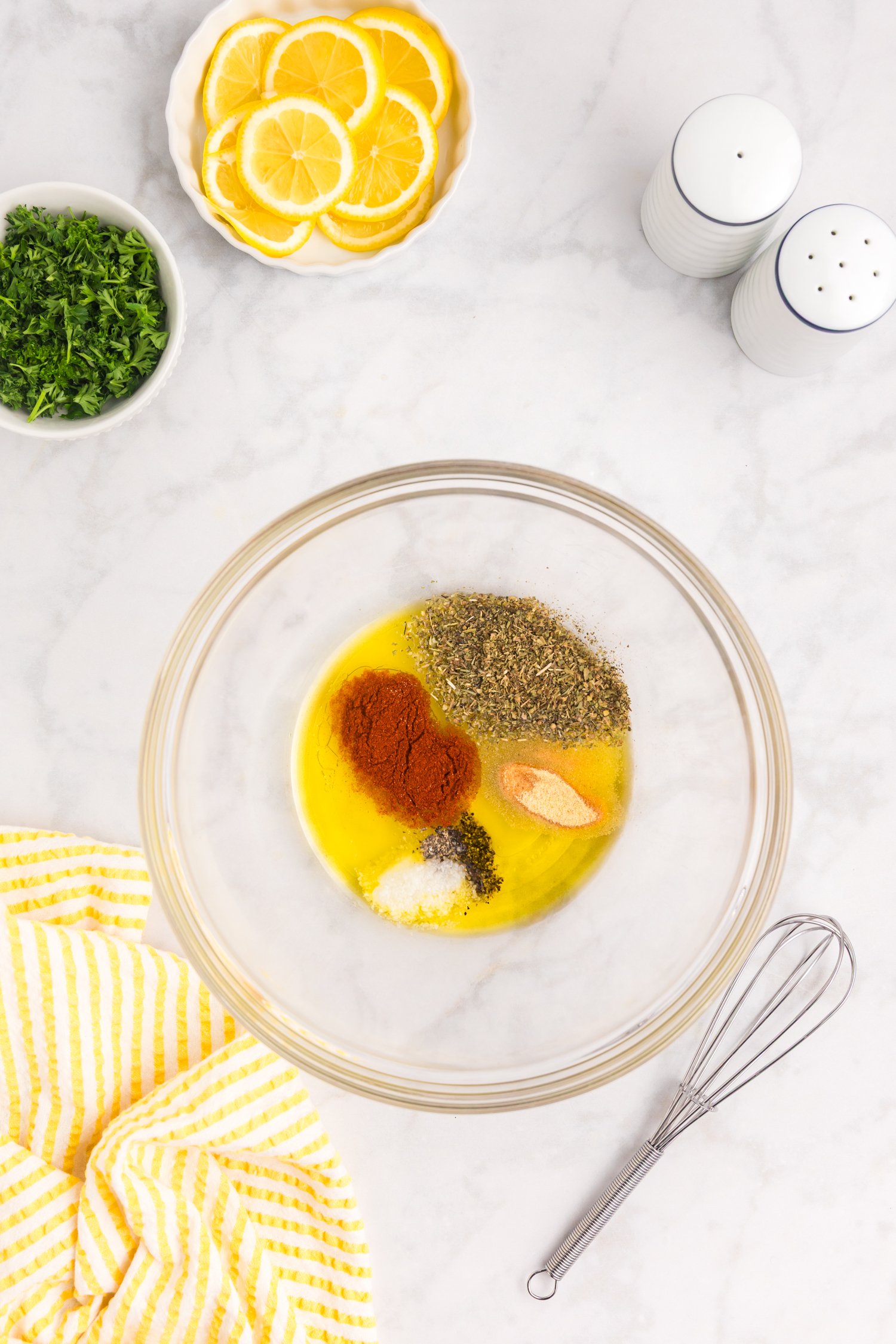 A glass bowl containing olive oil and various spices, including paprika, oregano, salt, pepper, and garlic powder, ready to be whisked together; lemon slices and parsley are placed above the bowl.