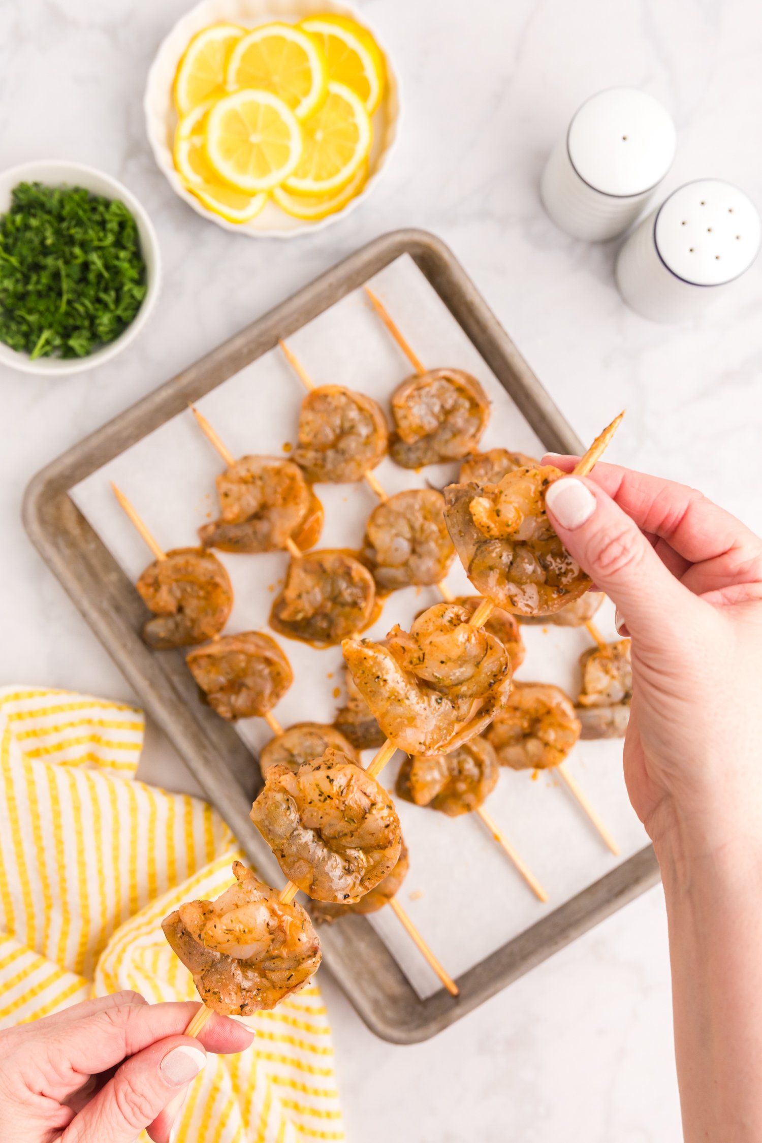 Hands are placing marinated shrimp onto skewers over a tray lined with parchment paper, preparing them for grilling.