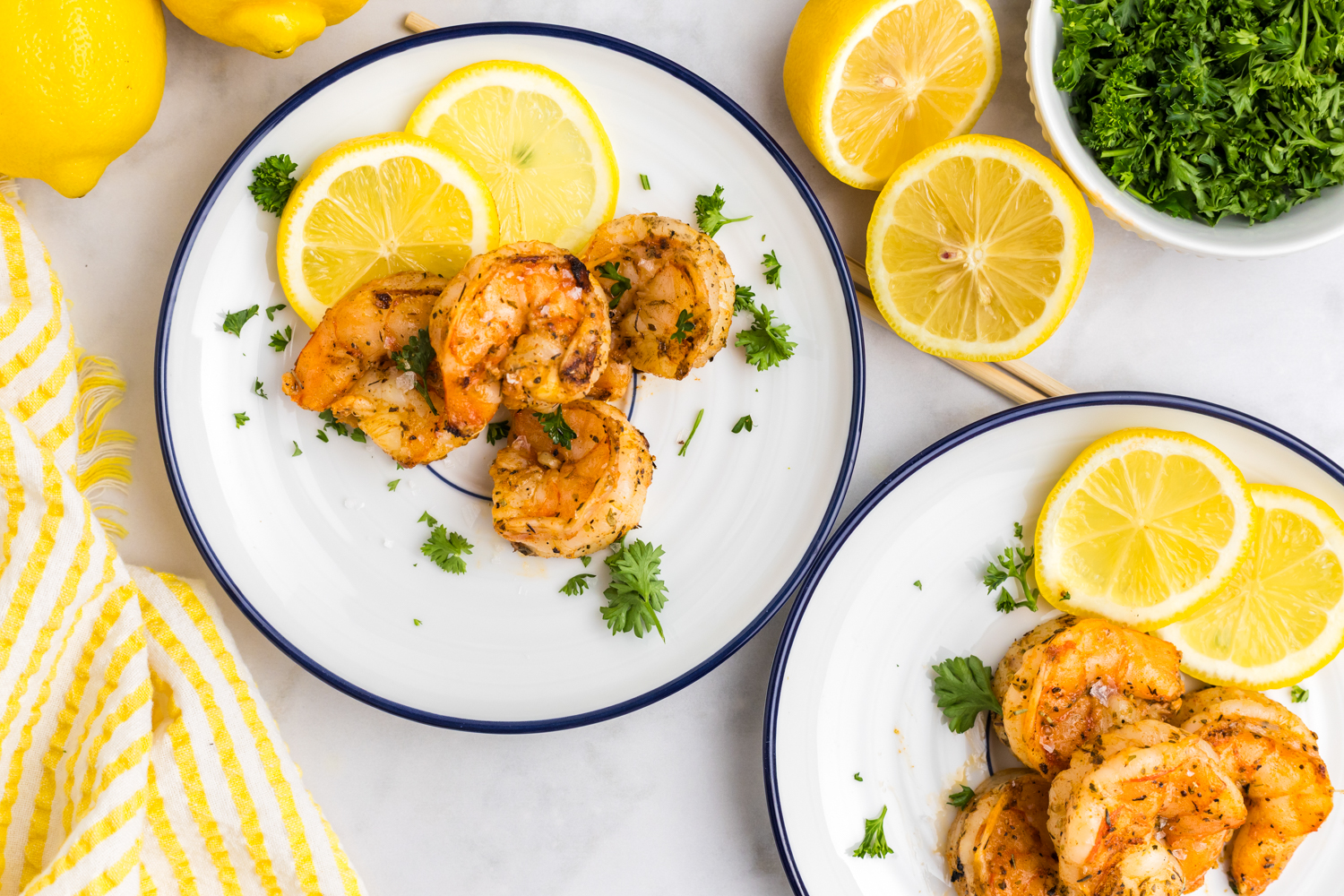 Overhead view of two white plates with blue trim, each containing a serving of grilled shrimp garnished with parsley and lemon slices; fresh parsley and halved lemons are scattered around for decoration.
