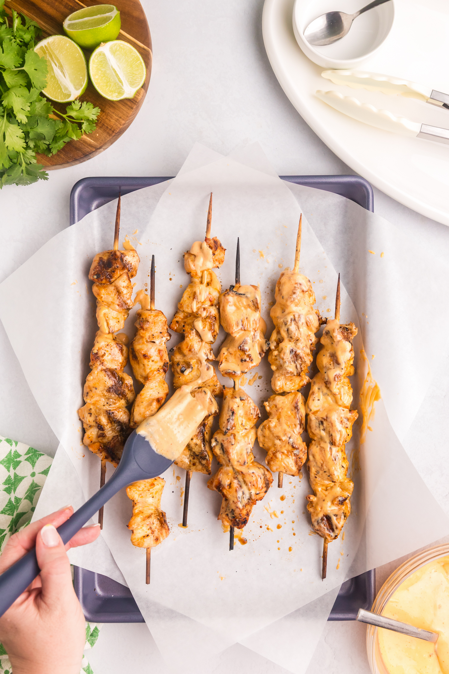 Grilled chicken skewers placed on parchment paper-lined tray being brushed with creamy Bang Bang sauce using a silicone brush.