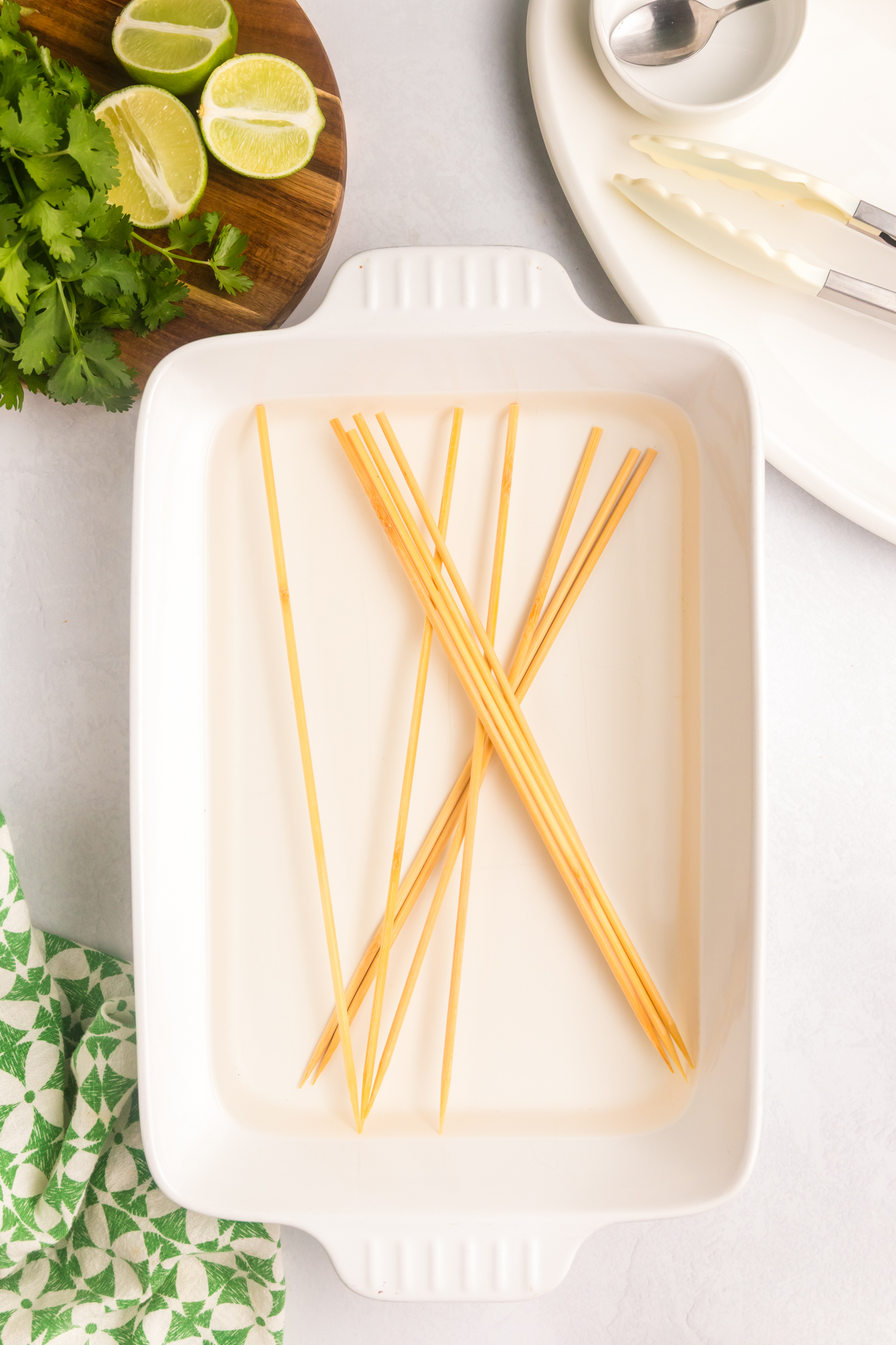 Bamboo skewers soaking in a white ceramic dish, placed next to halved limes, fresh cilantro, and kitchen utensils.