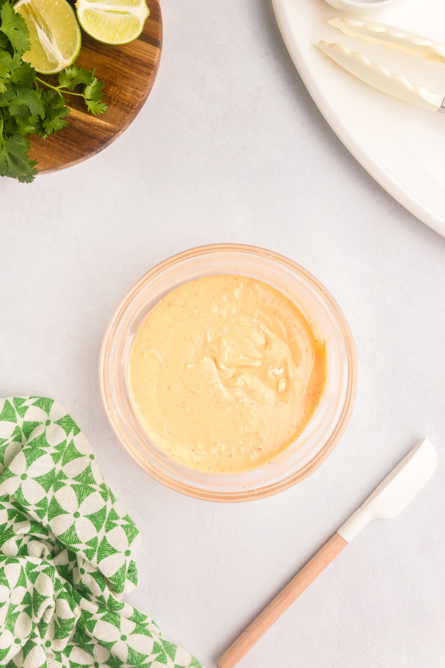 Smooth and creamy Bang Bang sauce blended in a glass bowl, with a rubber spatula and green patterned towel nearby.