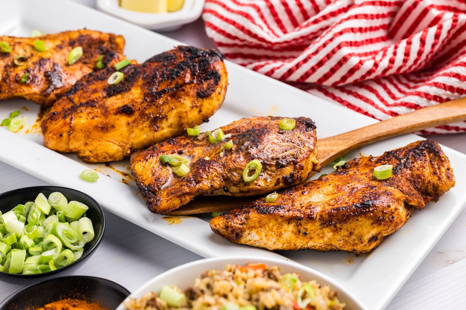 Cooked blackened chicken breasts garnished with chopped green onions served on a rectangular white platter, with a side of dirty rice and green onions.