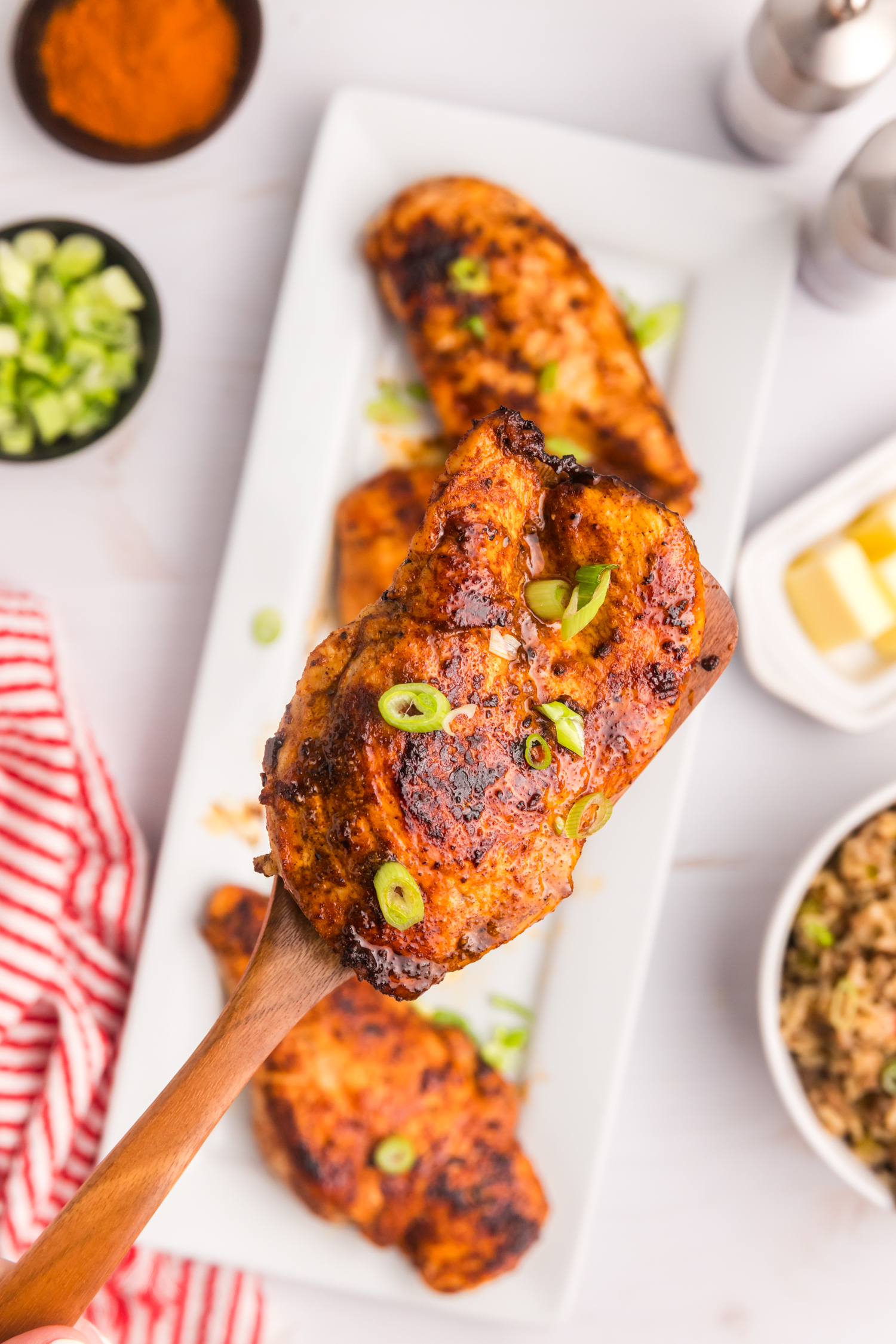 A close-up of a single cooked blackened chicken breast lifted from a serving tray with a wooden spatula, garnished with green onions.