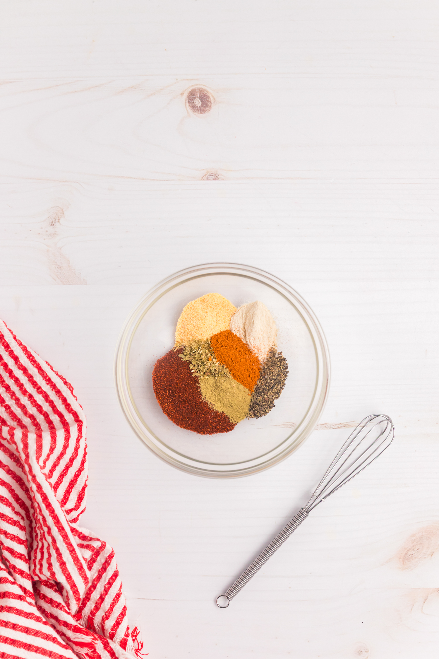 A bowl of assorted dry spices including paprika, garlic powder, onion powder, black pepper, oregano, and cayenne pepper, ready to be mixed with a whisk beside it.