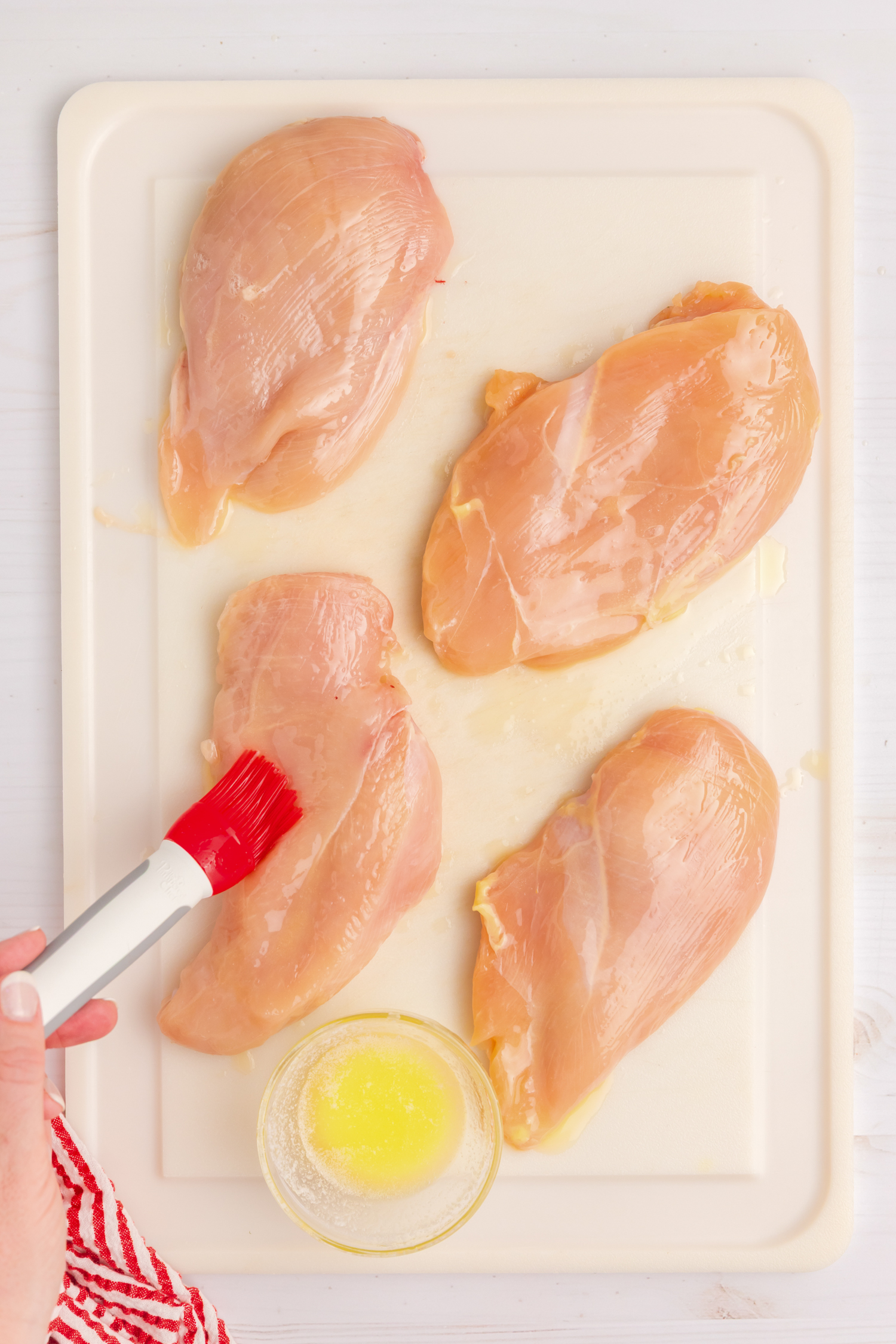 Four raw chicken breasts on a cutting board being brushed with melted butter using a red-tipped brush.