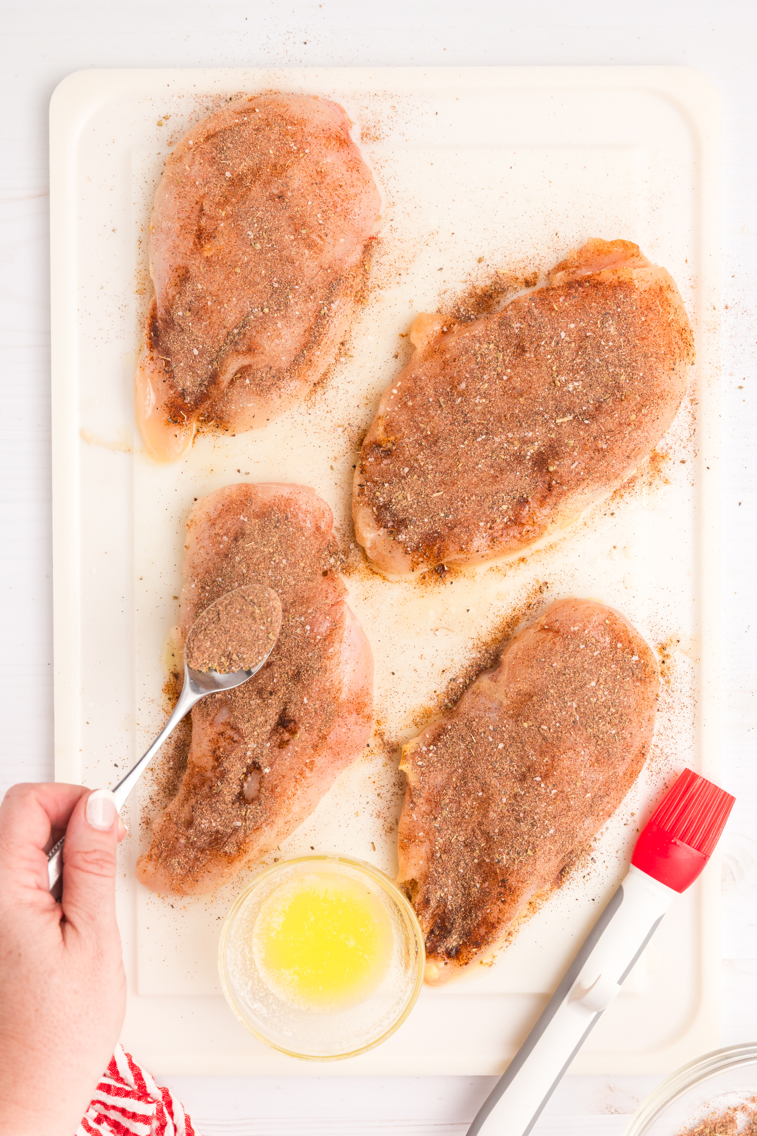 Raw chicken breasts being sprinkled with homemade blackened seasoning, with a bowl of melted butter and a basting brush nearby