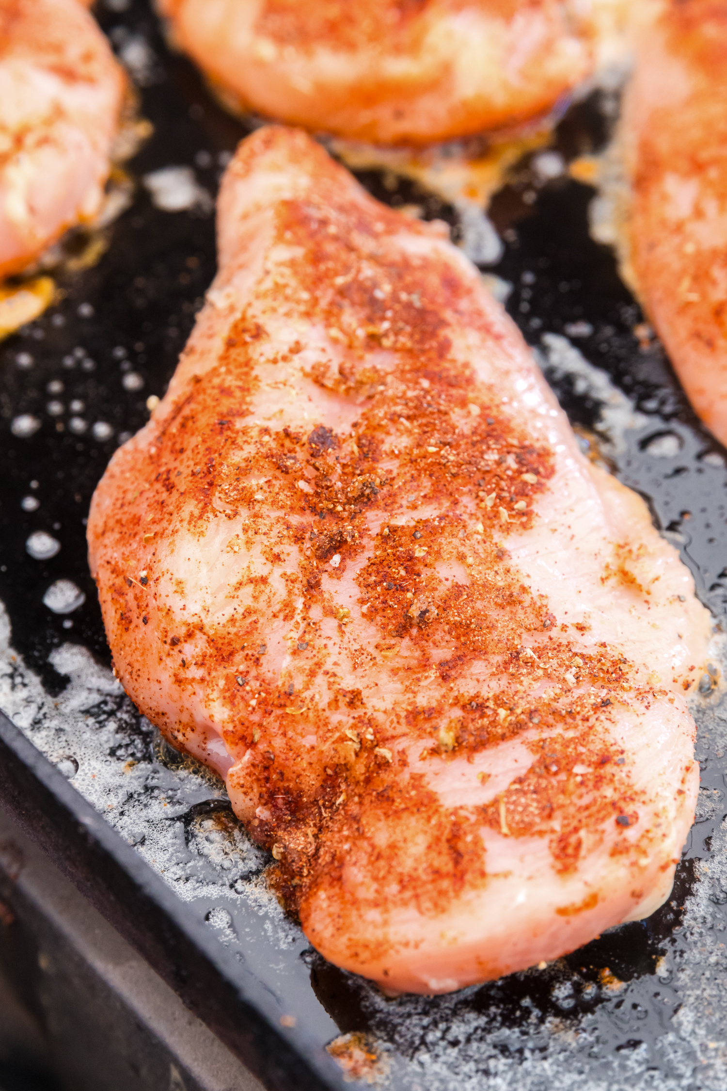 Raw chicken breasts coated in spices cooking on a greased flat-top griddle, starting to sear."