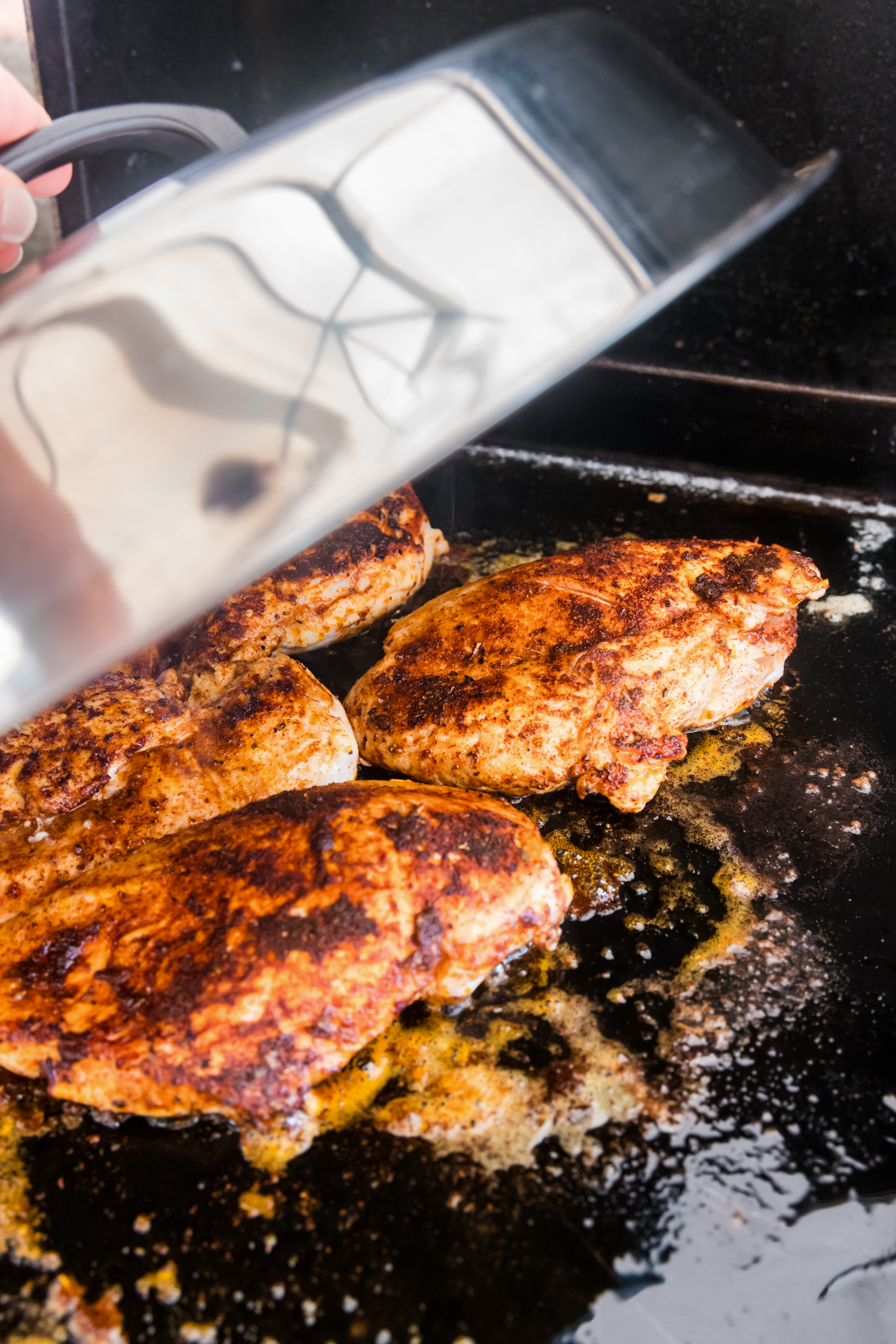 A lid being placed over blackened chicken breasts cooking on a griddle to trap heat and ensure thorough cooking.