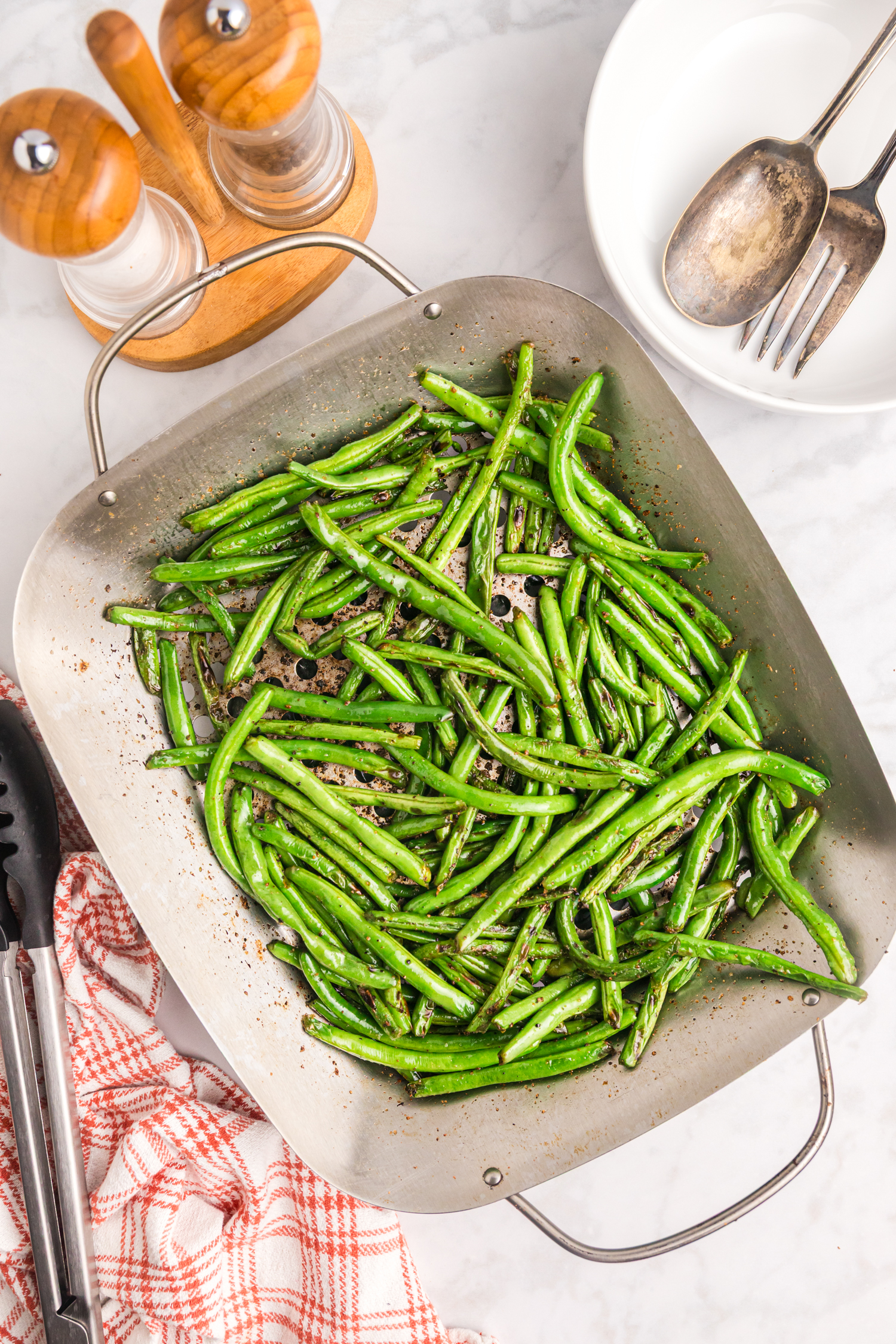 A large grilling basket of green beans on a kitchen counter, next to utensils and a red checkered towel, ready to be served.