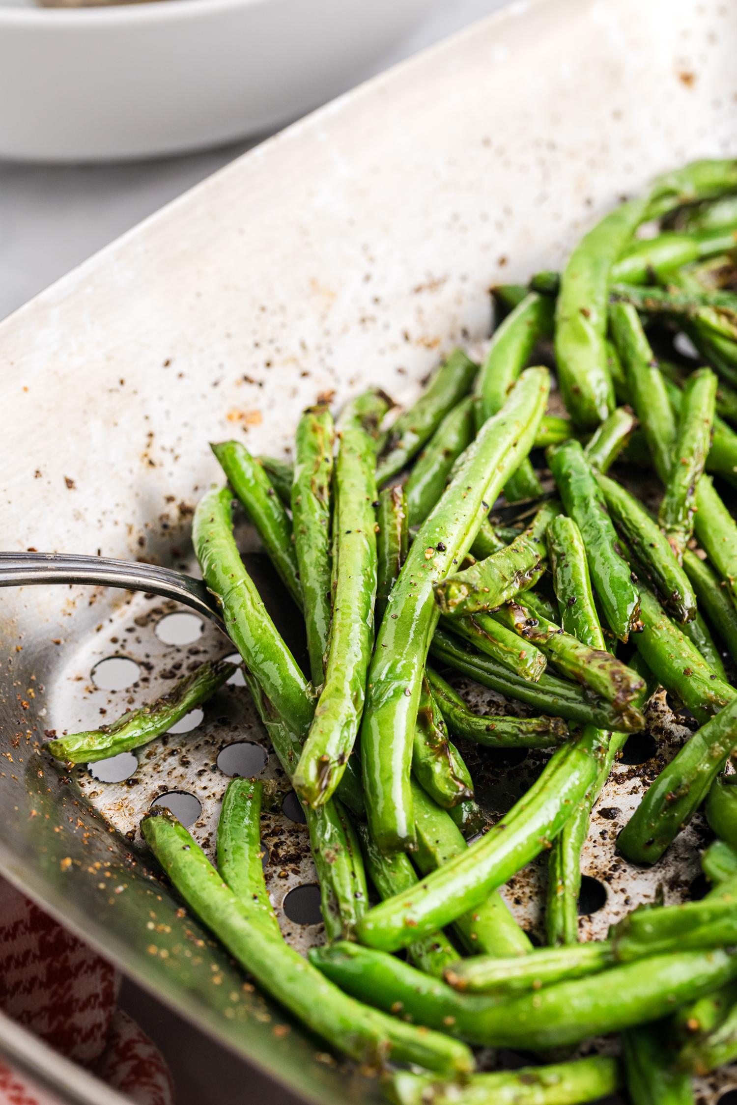 Close-up of grilled green beans in a grill basket, seasoned and charred, being stirred or served with a metal spoon.