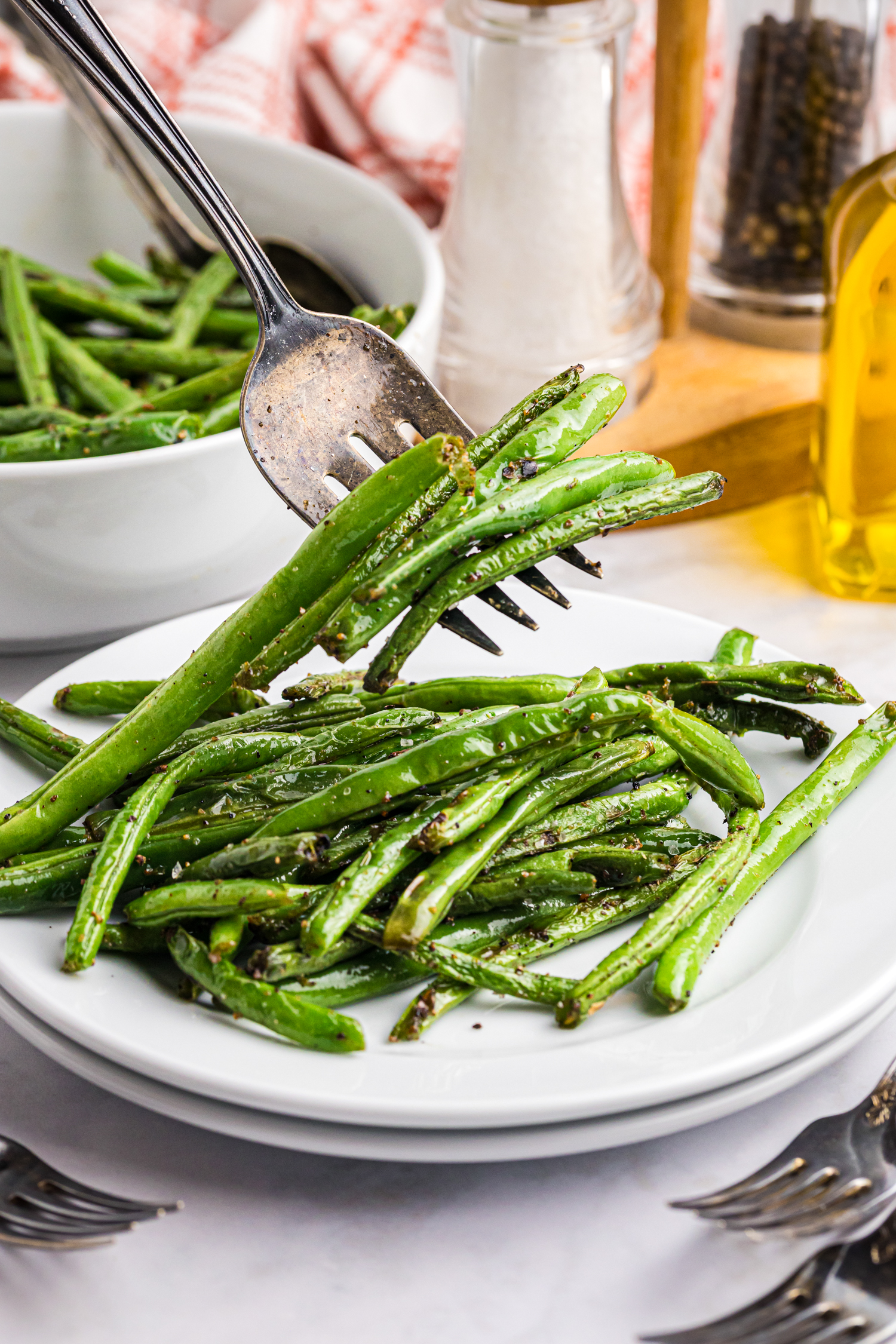 A fork lifting a portion of seasoned and grilled green beans from a white plate, with another serving bowl and pepper and salt shakers in the background.

