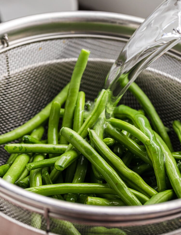 Fresh green beans being rinsed with water in a metal colander placed over a bowl or sink.