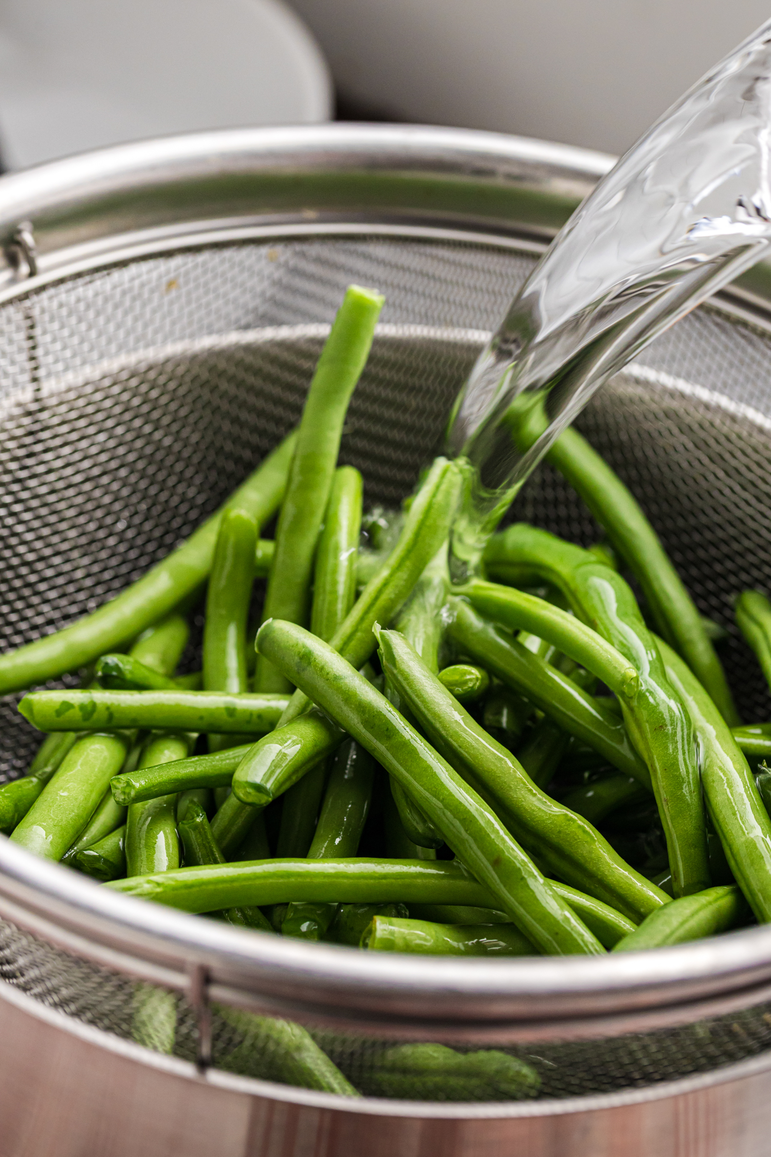 Fresh green beans being rinsed with water in a metal colander placed over a bowl or sink.