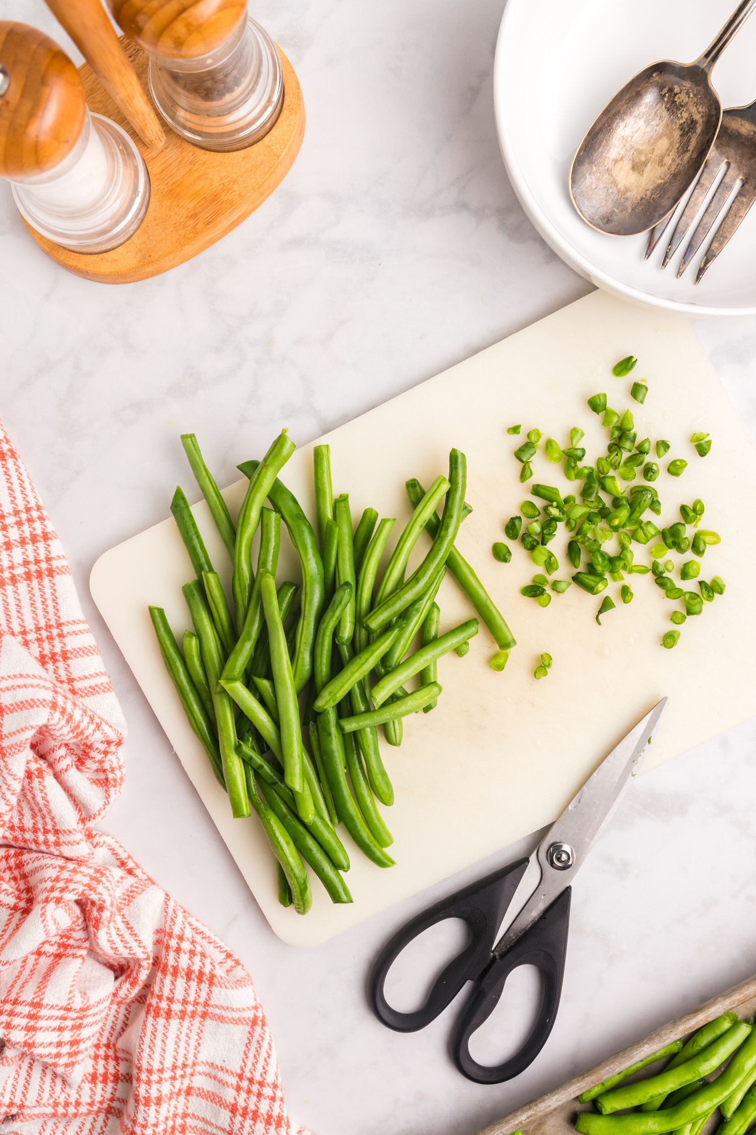 Green beans on a white cutting board, some chopped into small pieces with kitchen scissors, beside a red checkered cloth.