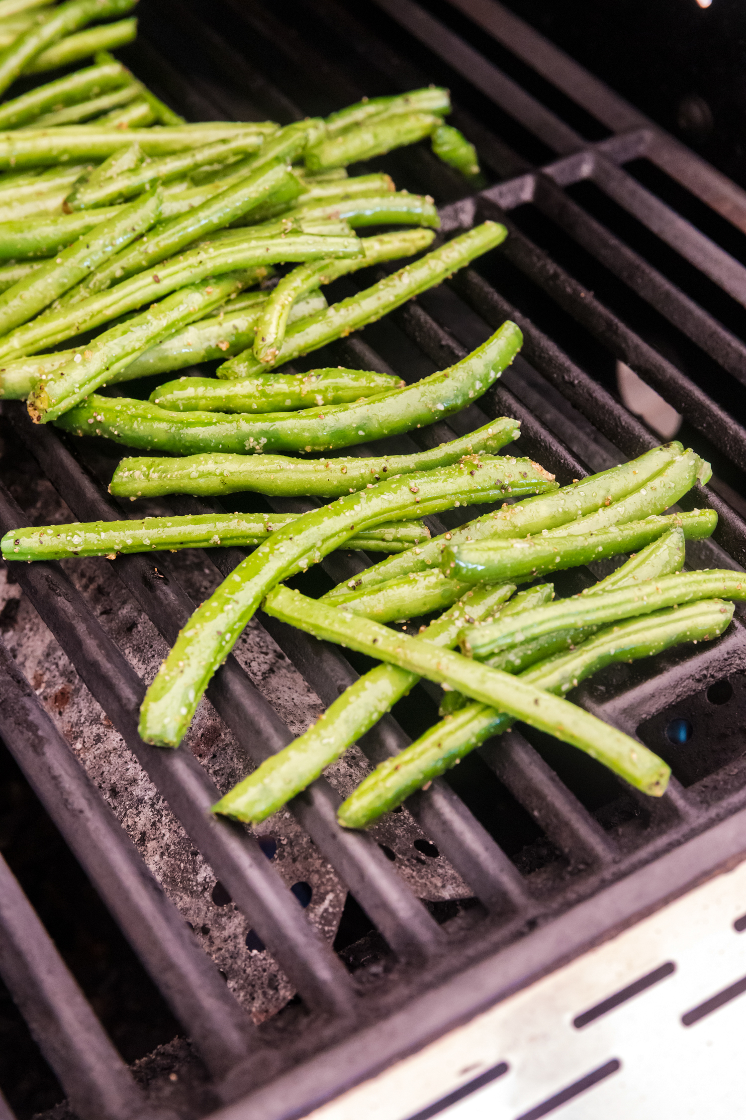 Green beans being grilled directly on a barbecue grate, lightly seasoned and starting to char.