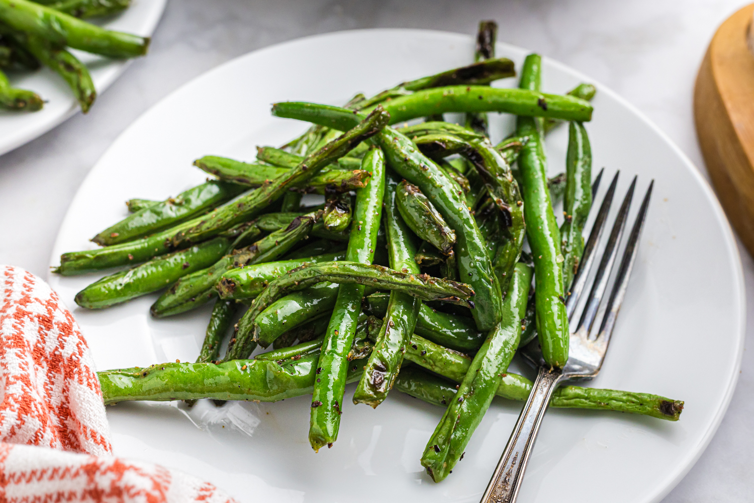 A close-up of grilled green beans served on a white plate with a silver fork, featuring lightly charred edges and a glossy seasoning. A red and white checkered napkin is partially visible on the side.








