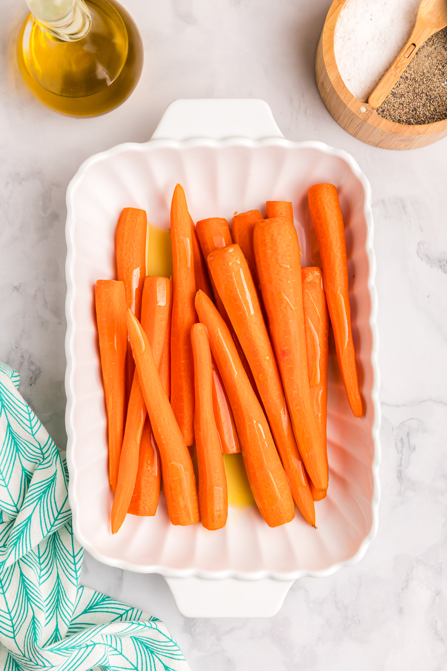Peeled whole carrots placed in a white scalloped ceramic baking dish with a drizzle of olive oil.