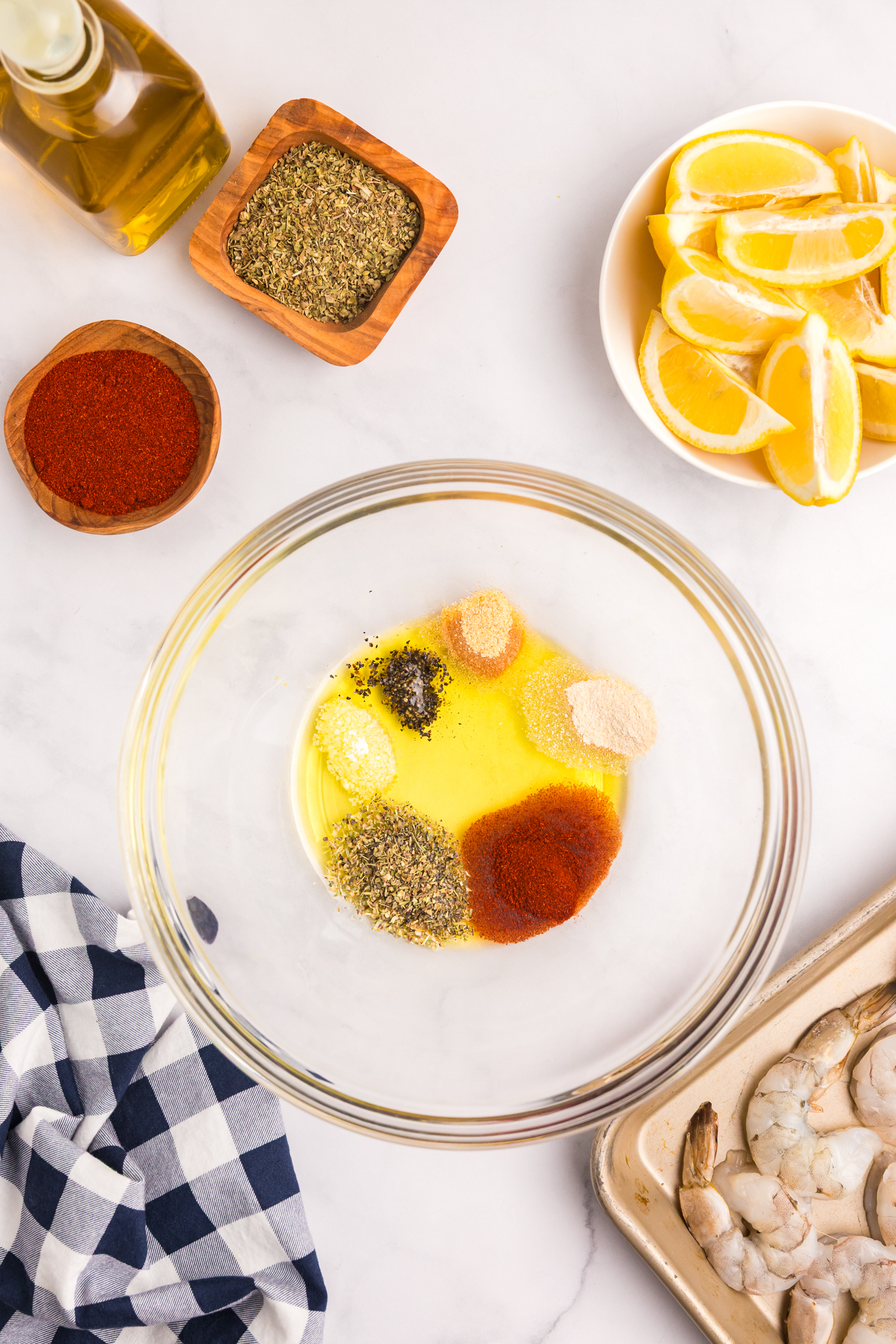 A glass mixing bowl containing olive oil and a variety of spices including paprika, oregano, onion powder, garlic powder, salt, and pepper. Ingredients are arranged around the bowl including lemon wedges, olive oil, and raw shrimp on a tray.
