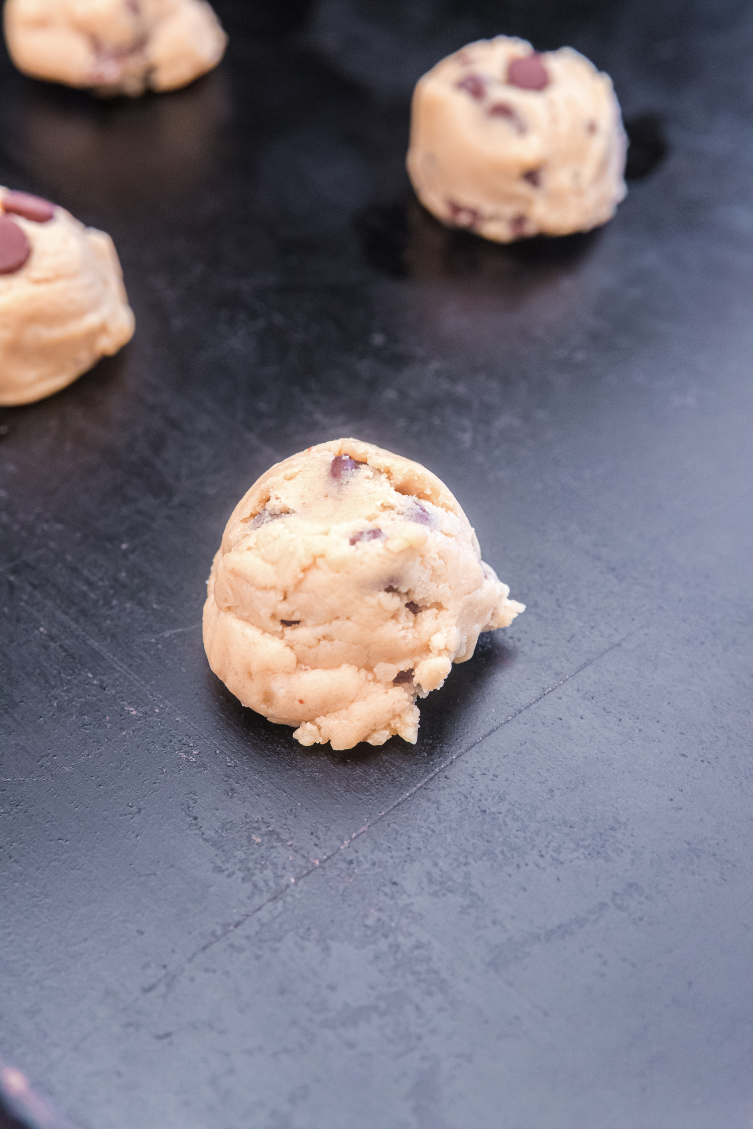 A close-up of a single raw chocolate chip cookie dough ball placed on a dark baking sheet.