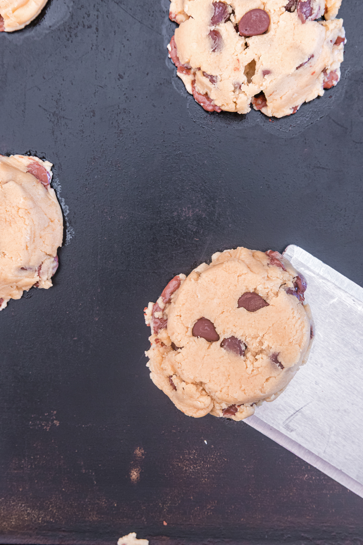 A spatula lifting a baked chocolate chip cookie from a dark baking sheet. The cookie is golden brown and studded with chocolate chips.