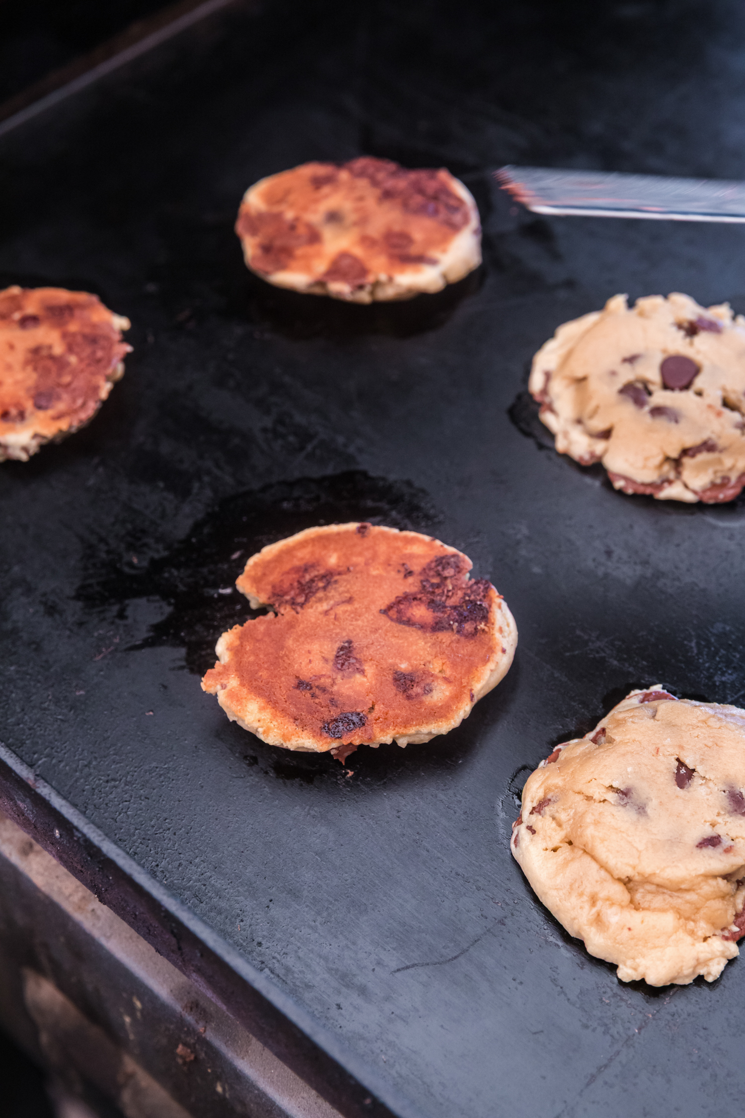 Chocolate chip cookie dough patties cooking on a Blackstone griddle. Some cookies have been flipped, showing a golden-brown, slightly crispy surface, while others are still raw on top.