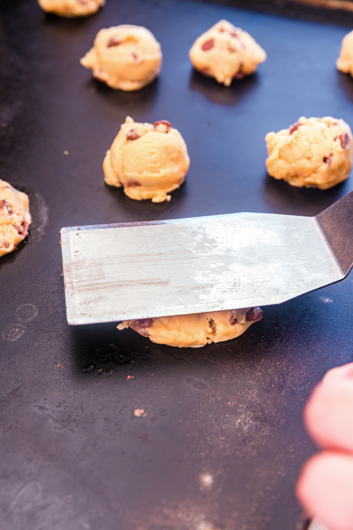 A metal spatula pressing down a ball of cookie dough on a griddle, flattening it into a patty. Several other dough balls are visible in the background.