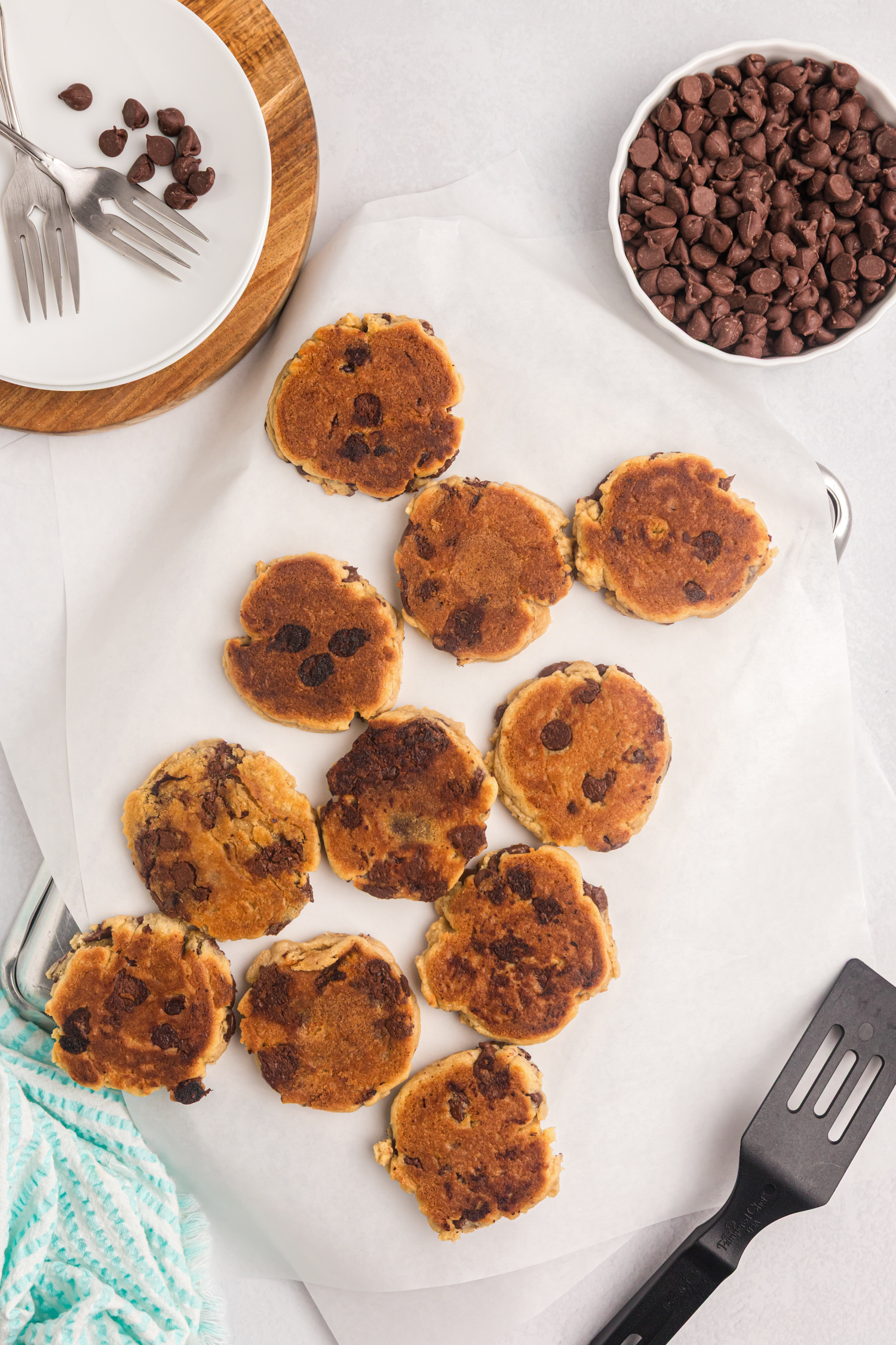 A dozen chocolate chip cookies with crisp, browned tops resting on white parchment paper. A bowl of chocolate chips, utensils, and a spatula surround the cookies on a clean white surface.