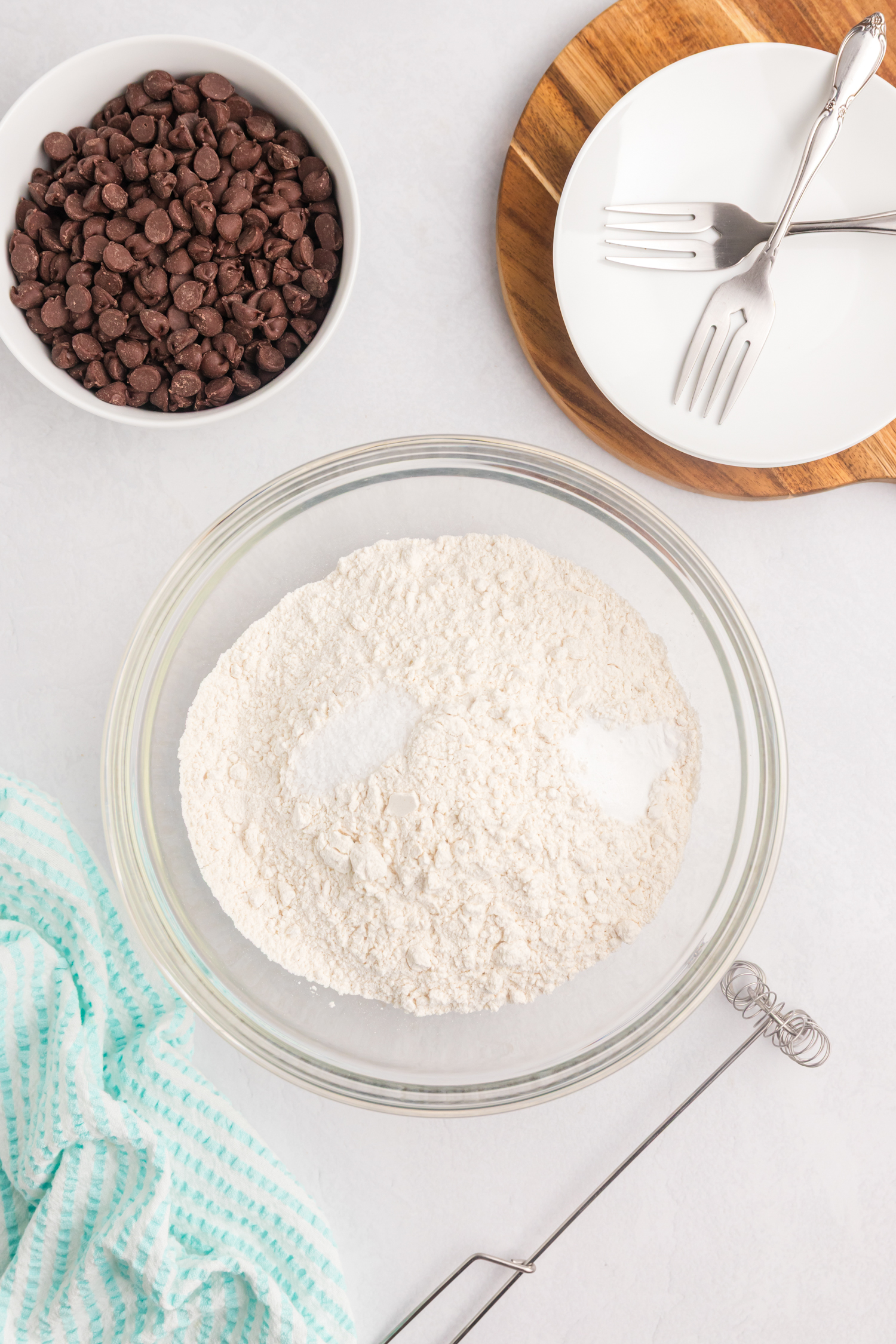 A glass mixing bowl filled with flour, baking soda, and salt, placed on a white surface. Surrounding the bowl are a whisk, a bowl of chocolate chips, a striped kitchen towel, and stacked white plates with silver forks on a wooden board.