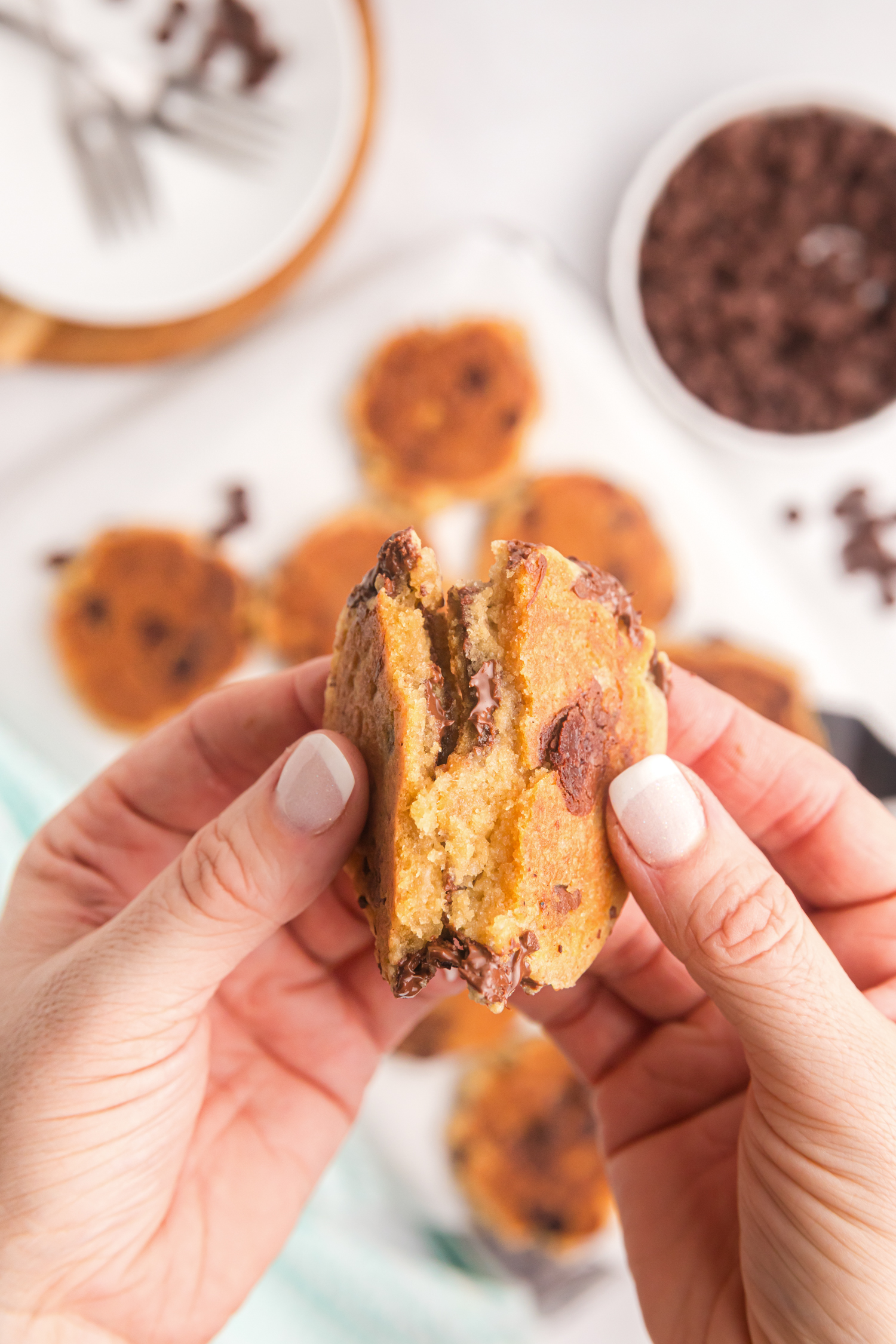 A pair of hands pulling apart a freshly cooked chocolate chip cookie, revealing a warm, gooey center with melted chocolate chips.

