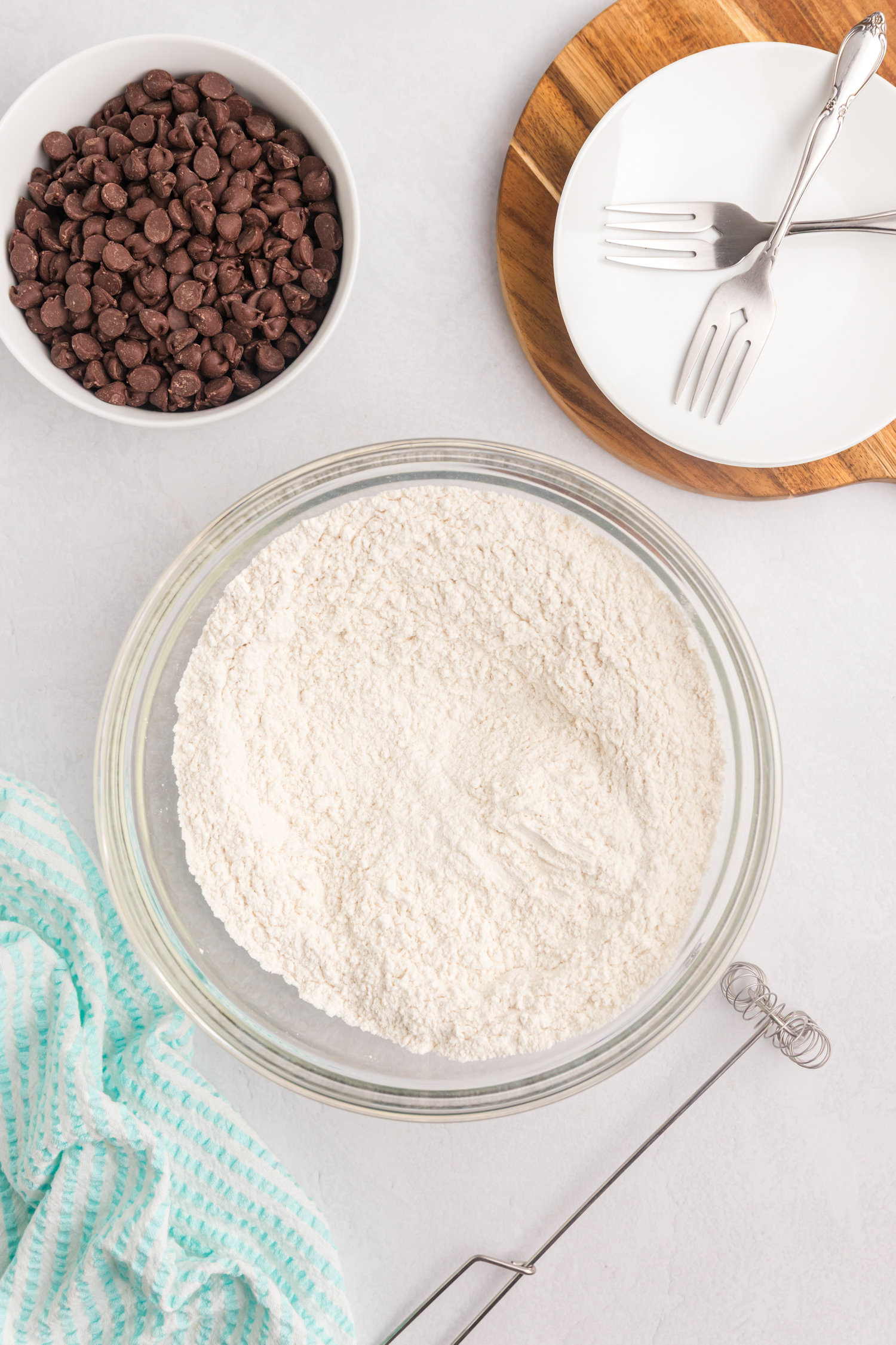 The dry ingredients (flour, baking soda, and salt) have been fully mixed in the glass bowl. The same bowl of chocolate chips, plates, and utensils remain nearby.
