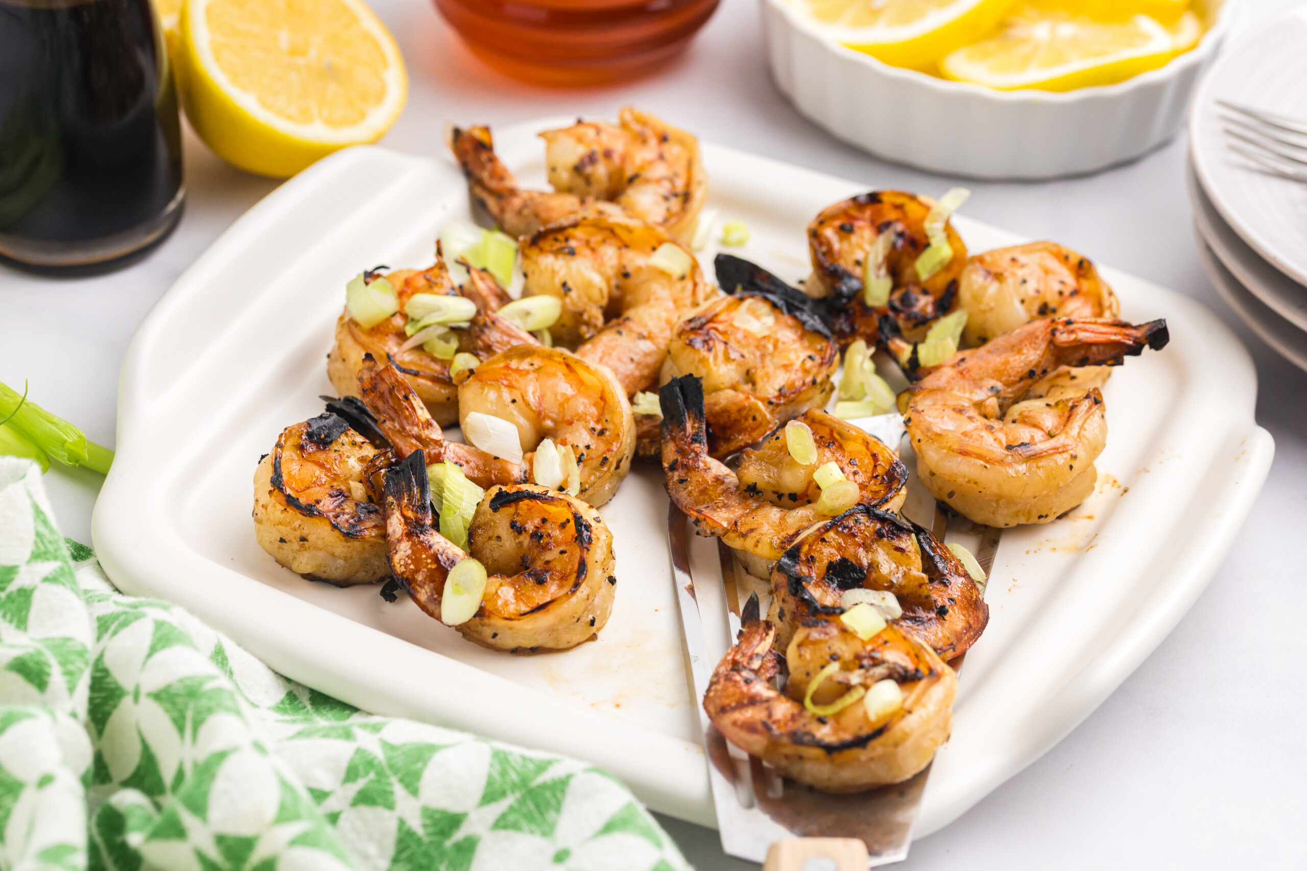 Finished grilled shrimp served on a white platter, skewered and topped with green onions and lemon wedges nearby.