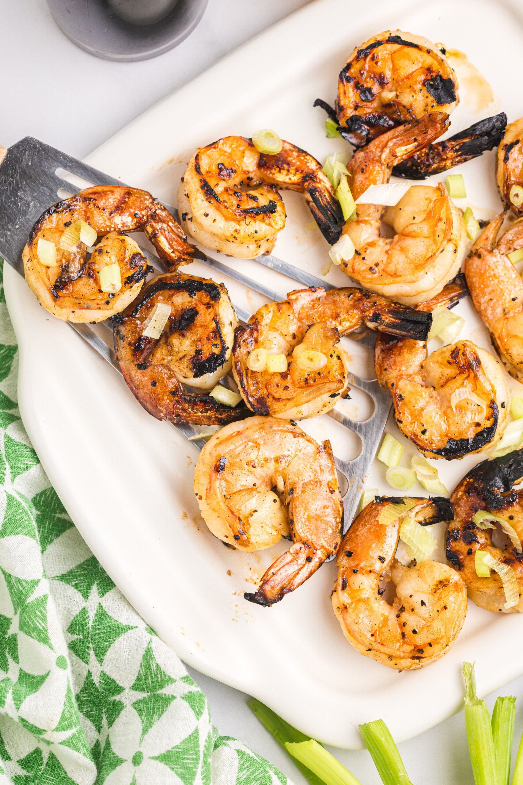 Close-up of grilled shrimp served on a white platter with metal skewers, garnished with chopped green onions and slightly charred edges, ready to eat.

