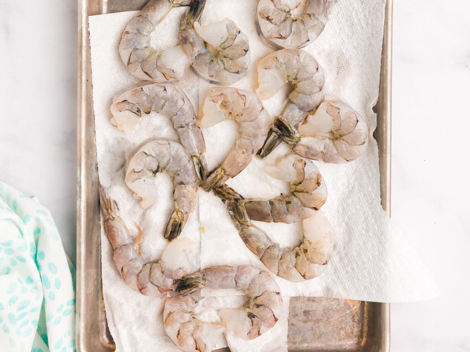 Raw, peeled shrimp laid out on a metal tray lined with paper towels, surrounded by lemon wedges, olive oil, seasoning mix, and salt and pepper shakers.