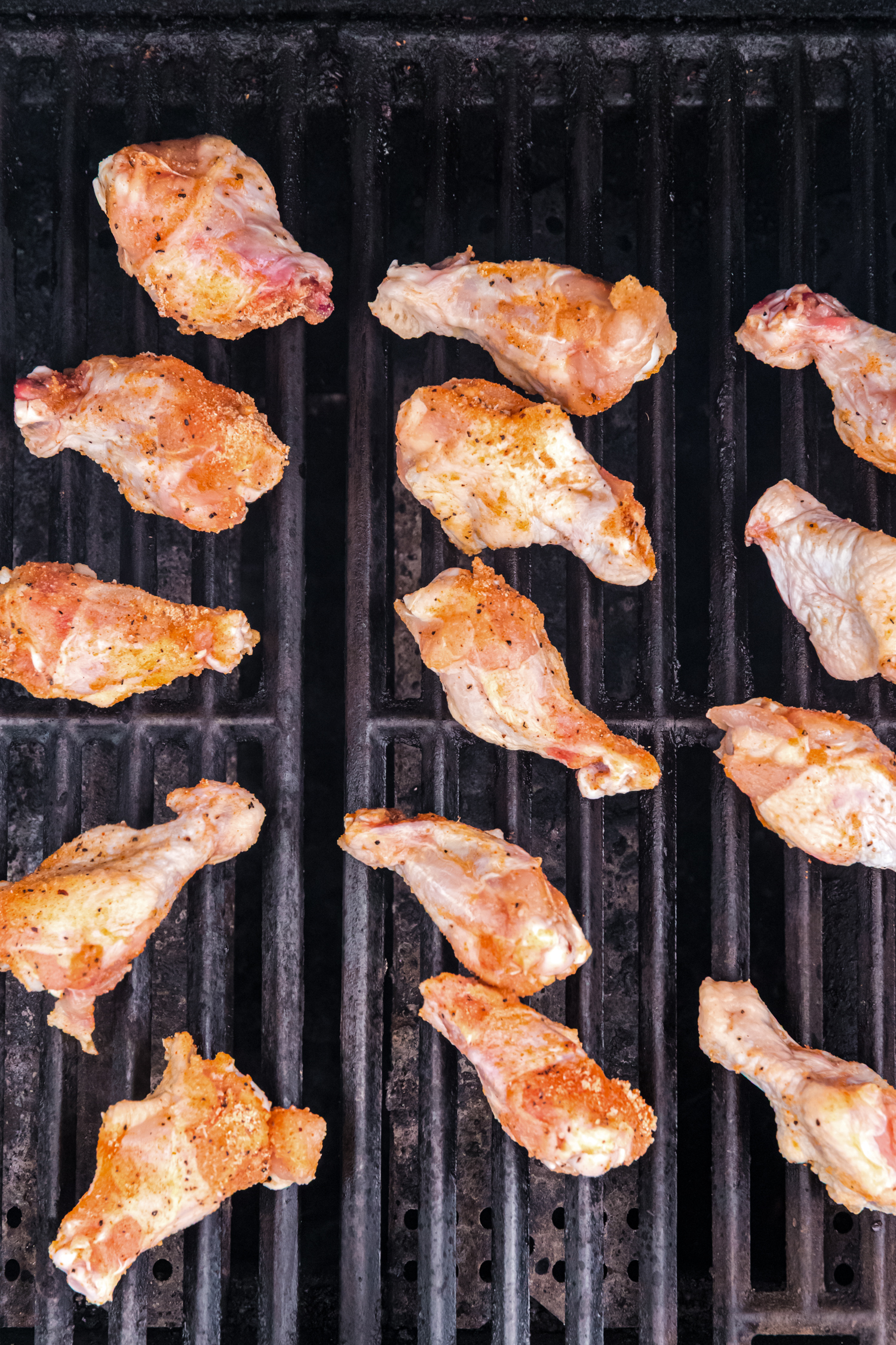 Seasoned raw chicken wings spread out on a grill, beginning to cook.