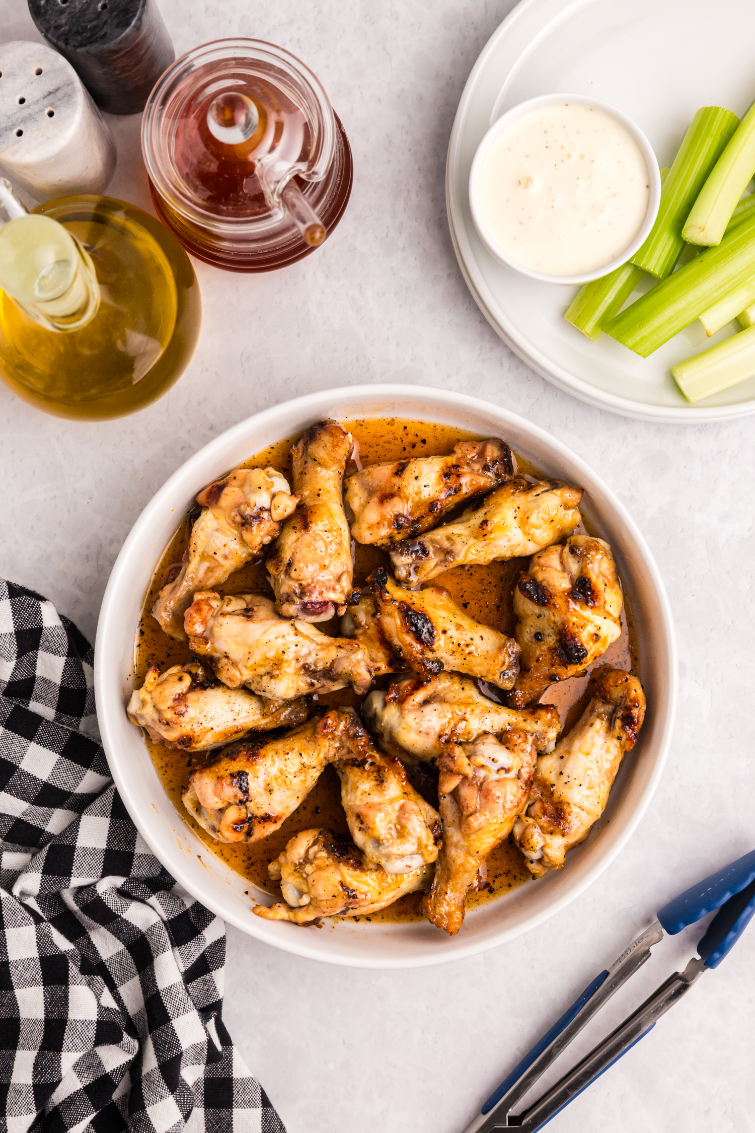 Bowl of grilled chicken wings coated in hot honey sauce, with celery sticks and dip on a plate nearby.