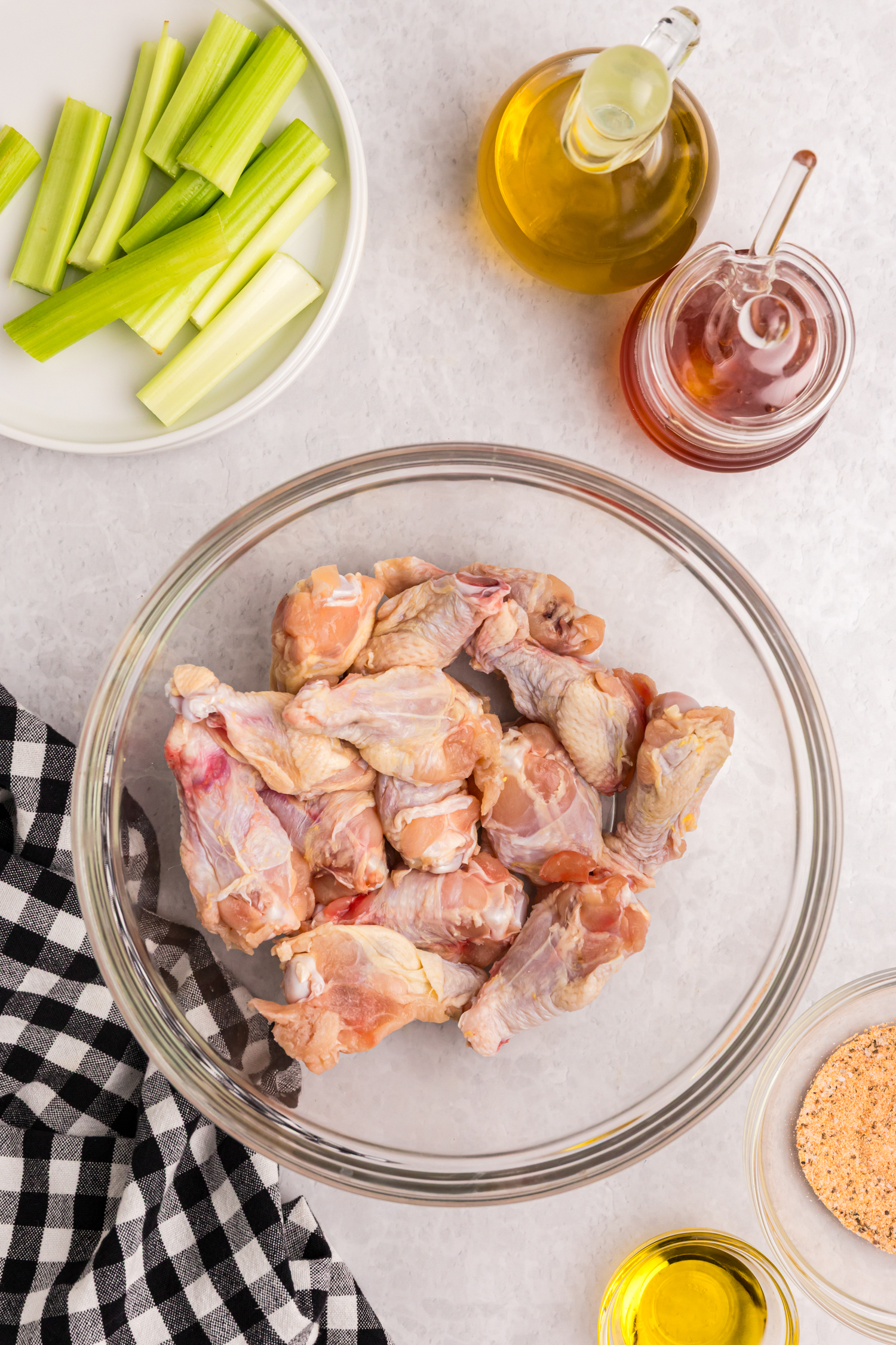 Raw chicken wings in a large glass bowl with celery sticks, oil, and honey jars nearby.