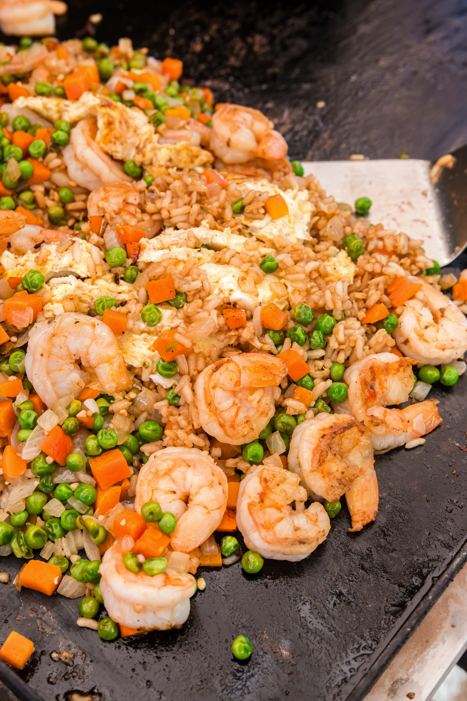 Fried rice with shrimp, peas, carrots, onions, and scrambled eggs being mixed together on a griddle.
