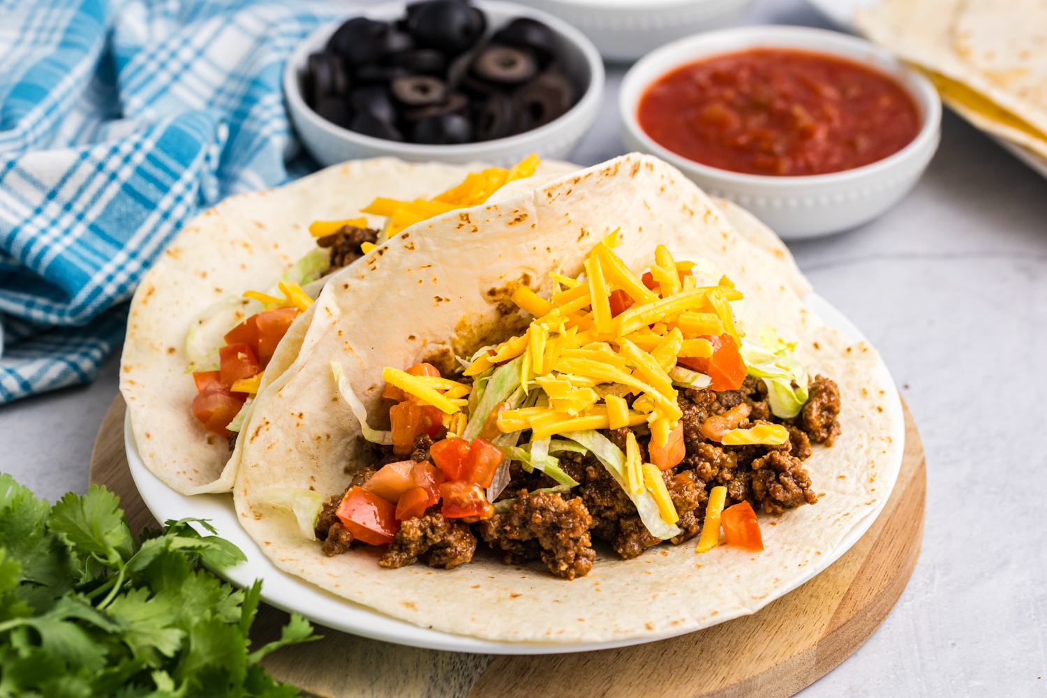 Two soft flour tortillas filled with seasoned ground beef, shredded lettuce, diced tomatoes, and shredded cheddar cheese, served on a white plate with bowls of black olives and salsa in the background.