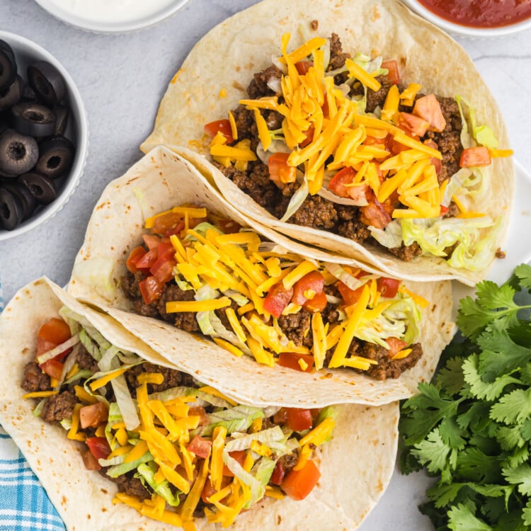 Three soft flour tacos filled with ground beef, lettuce, diced tomatoes, and cheddar cheese, displayed on a white surface with bowls of sour cream, salsa, and black olives nearby.