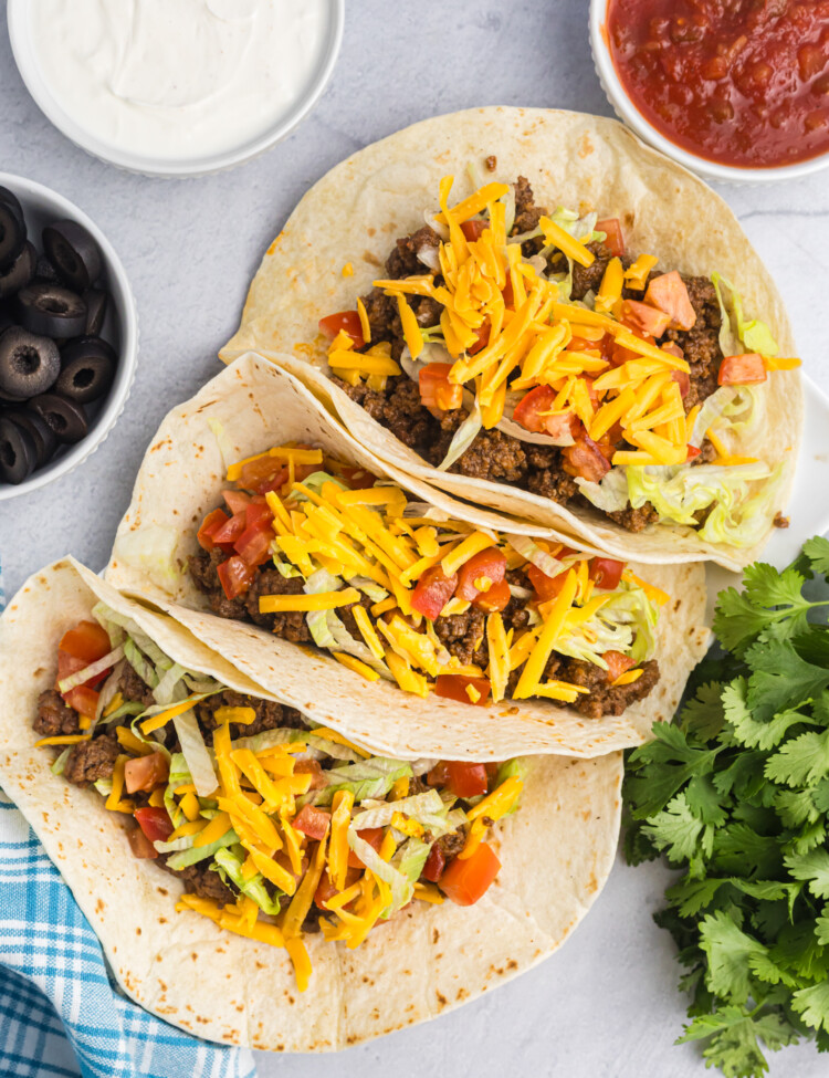 Three soft flour tacos filled with ground beef, lettuce, diced tomatoes, and cheddar cheese, displayed on a white surface with bowls of sour cream, salsa, and black olives nearby.