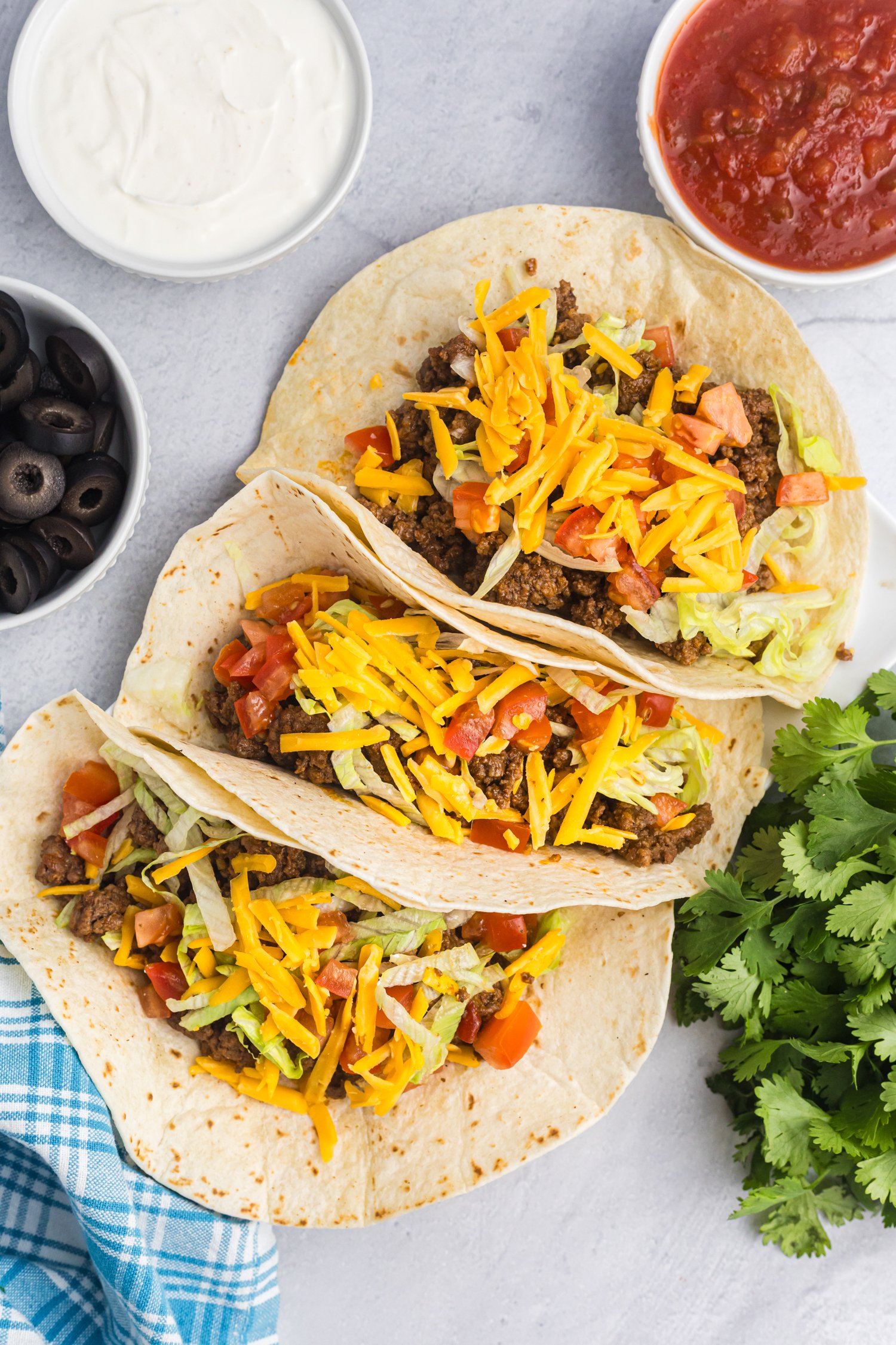Three soft flour tacos filled with ground beef, lettuce, diced tomatoes, and cheddar cheese, displayed on a white surface with bowls of sour cream, salsa, and black olives nearby.