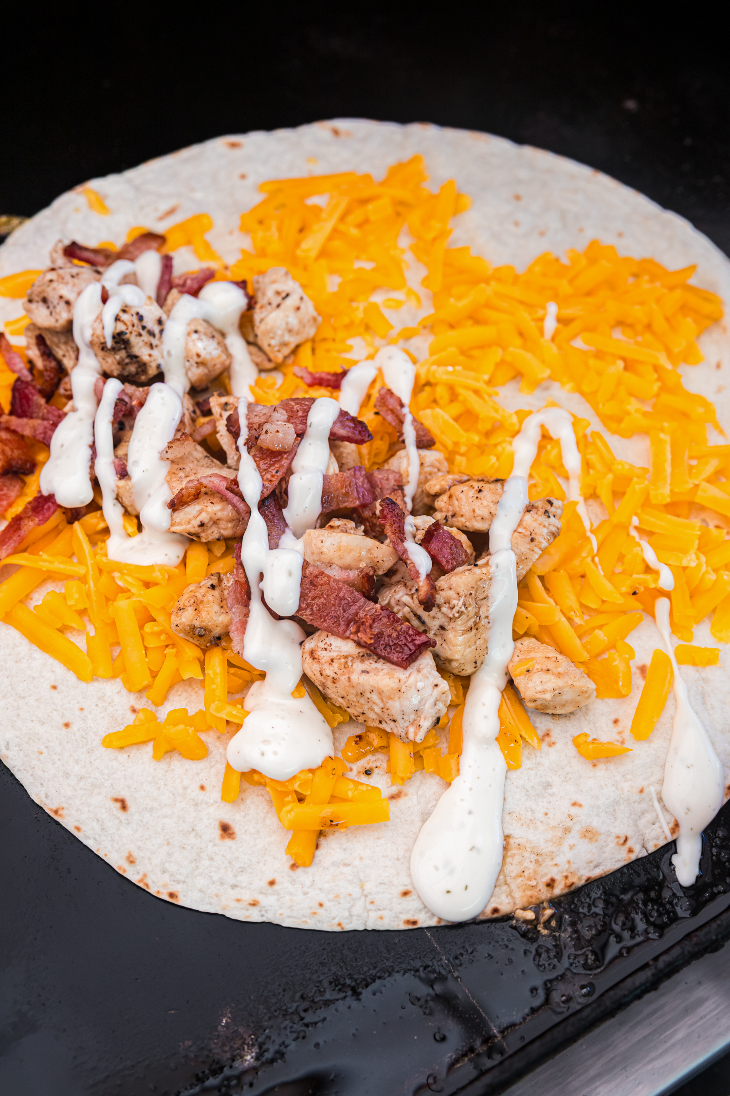 Tortilla on a griddle topped with shredded cheddar cheese, cooked chicken, crispy bacon, and ranch dressing.
